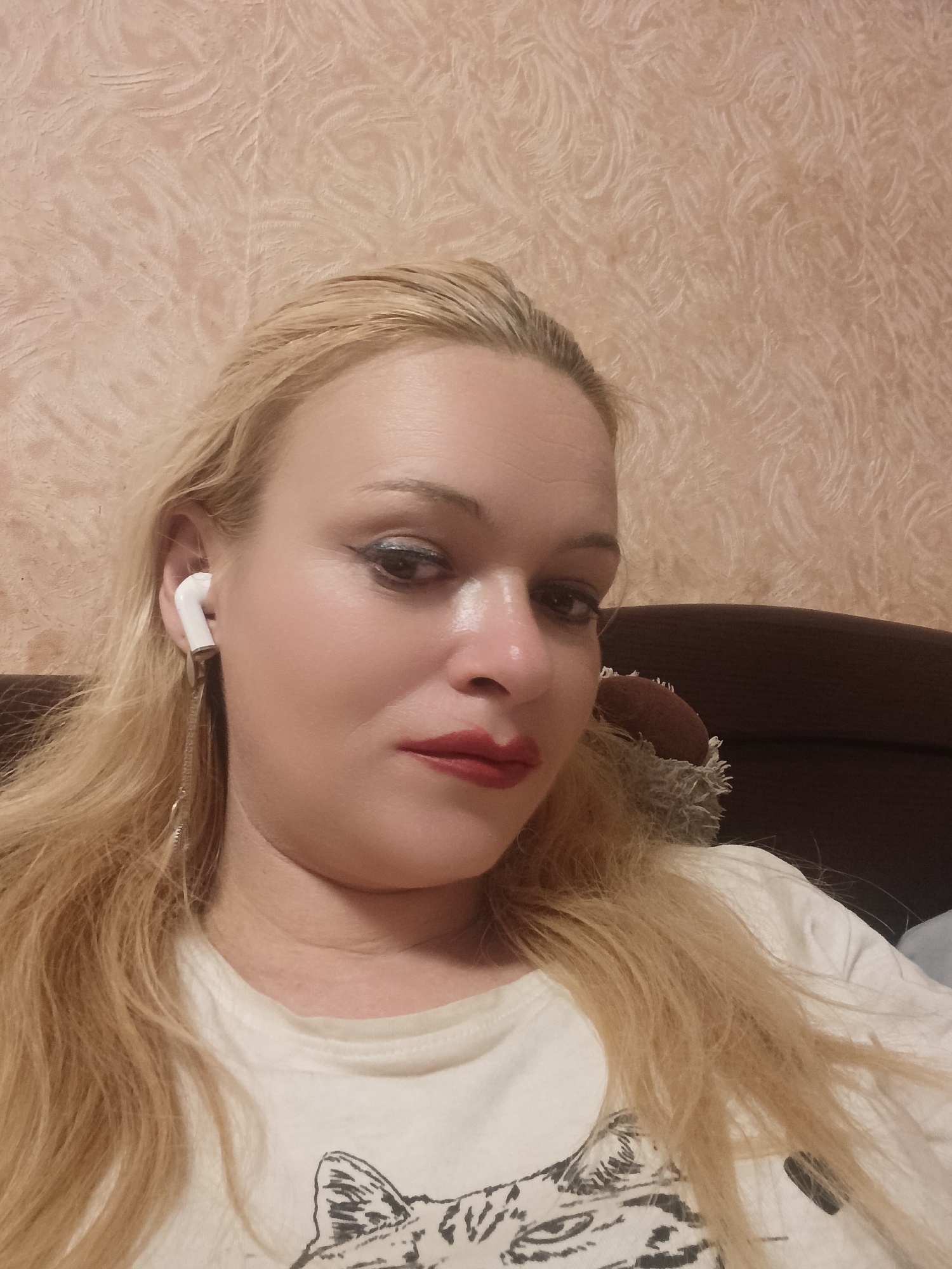 The image is a close-up, waist-up portrait of a blonde woman with fair skin, wearing a white t-shirt with a black graphic of a cat. She has red lipstick and dark eyeliner. A white earbud with a chain-like accessory dangles from her left ear. The background appears to be a textured wall. The lighting suggests it is indoors, possibly in the evening or at night. The woman is looking towards the camera with a neutral expression. There is no visible text in the image. The overall impression is a casual selfie taken indoors.