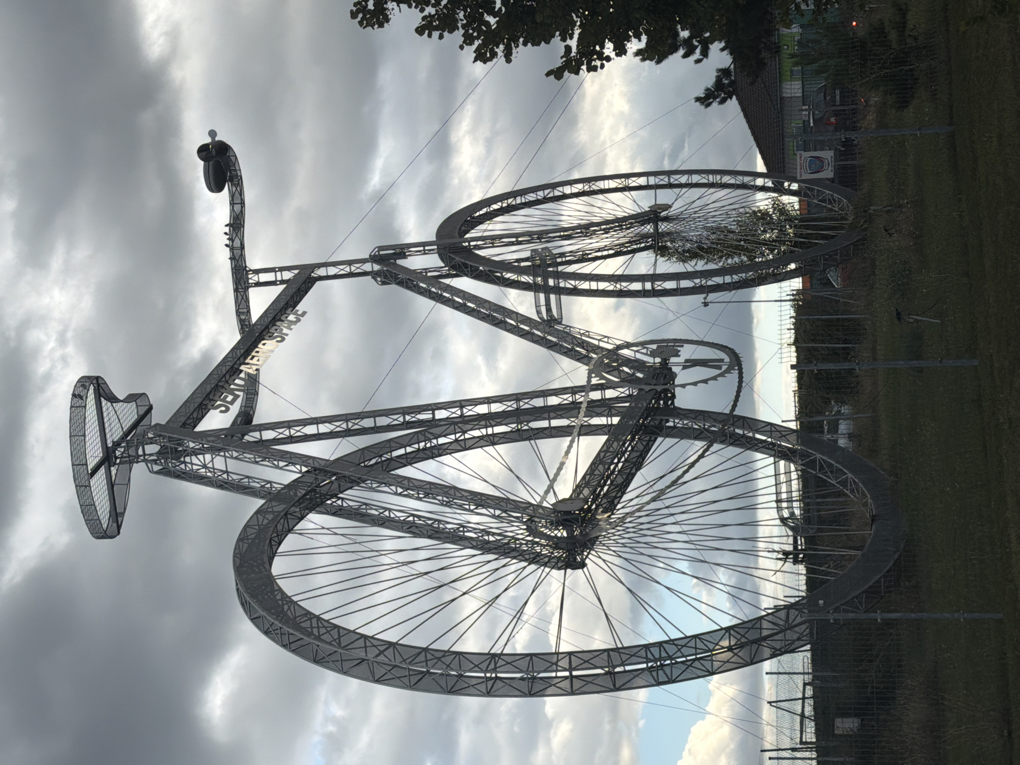 A large, gray, metal sculpture of a penny-farthing bicycle stands outdoors against a cloudy sky. The sculpture is located in Louny, Czechia. The sculpture's front wheel is significantly larger than its rear wheel. The text "SENTINEL AEROSPACE" is visible on the frame of the sculpture. A small propeller-like object is attached to the handlebars. Several birds are perched on the handlebars. In the background, there is a building with a dark roof, trees, a fence, and a grassy area. A car is visible behind the fence near the building.