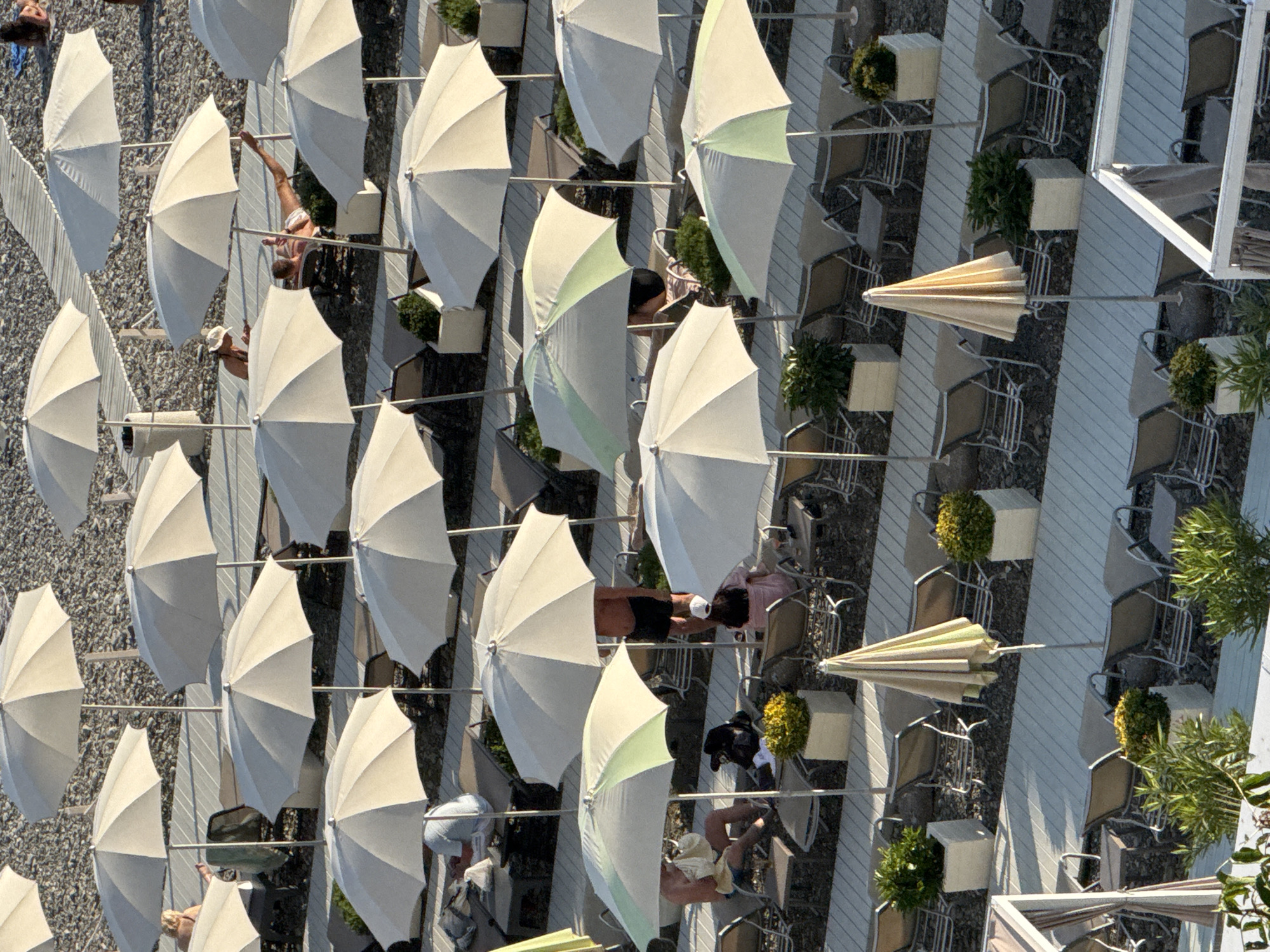 The aerial view captures a sun-drenched beach resort in Sochi, Russia, characterized by rows of unoccupied lounge chairs and numerous open and closed beach umbrellas. The beach itself is a stony expanse, typical of the Black Sea coast. The organized arrangement of lounge chairs and umbrellas suggests a well-maintained tourist facility. The time of day appears to be late afternoon, indicated by the long shadows cast by the umbrellas and the warm lighting. There are a few individuals present, scattered among the chairs and under the umbrellas, some appearing to be relaxing or preparing to depart as the day winds down. The scene conveys a sense of leisure and tranquility associated with a beach vacation.
