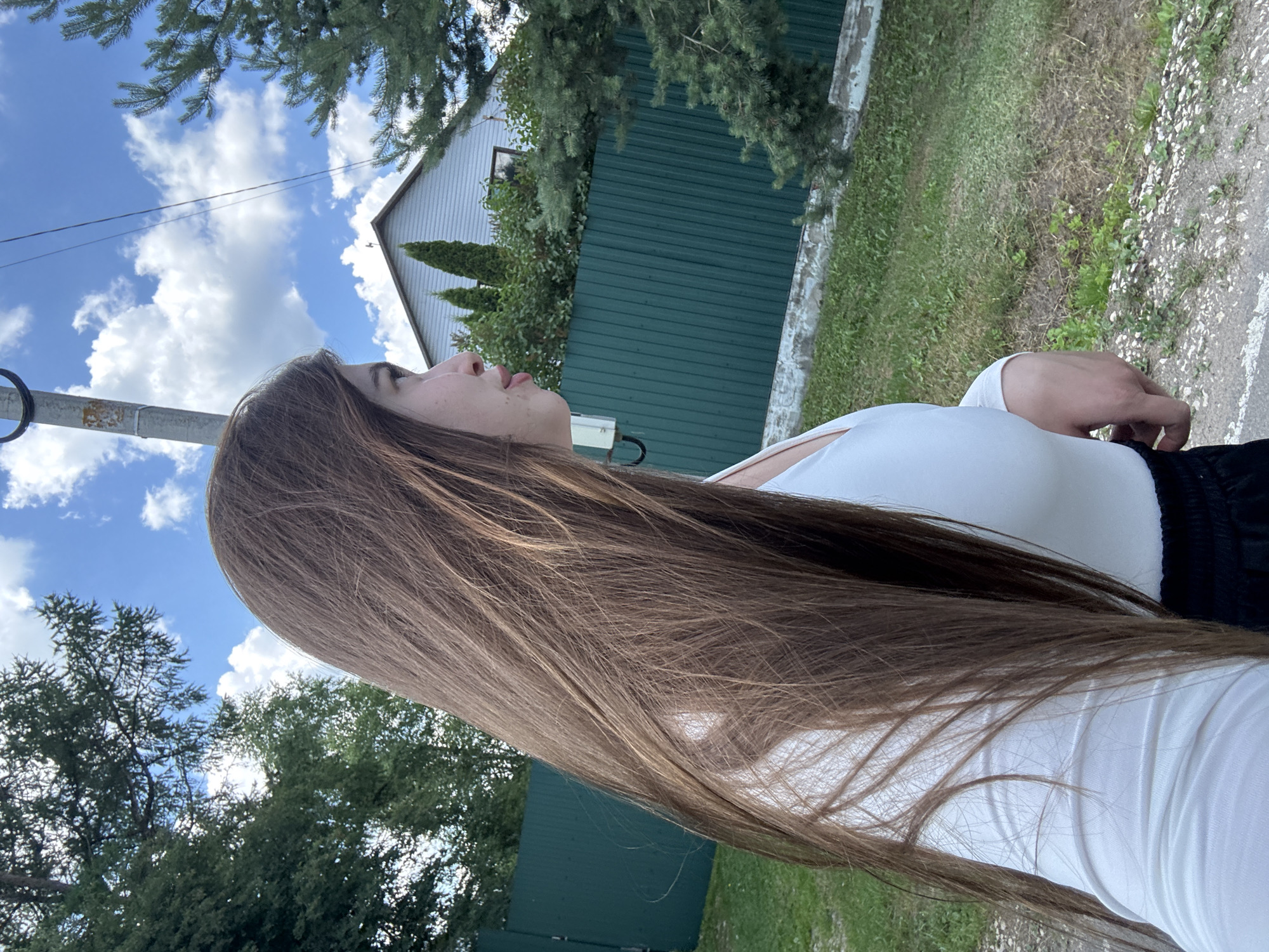 The image shows a young woman with long, brown hair, facing to the right. She is wearing a white long-sleeved shirt. In the background, there is a green metal fence, some trees, a house with a grey roof, and a blue sky with white clouds. The scene appears to be in a rural or suburban area. The woman is standing on a paved surface, possibly a road or sidewalk, with grass and dirt to her right. The presence of a utility pole with wires suggests an urban or semi-urban setting. The location is stated to be Мещёры, Russia.