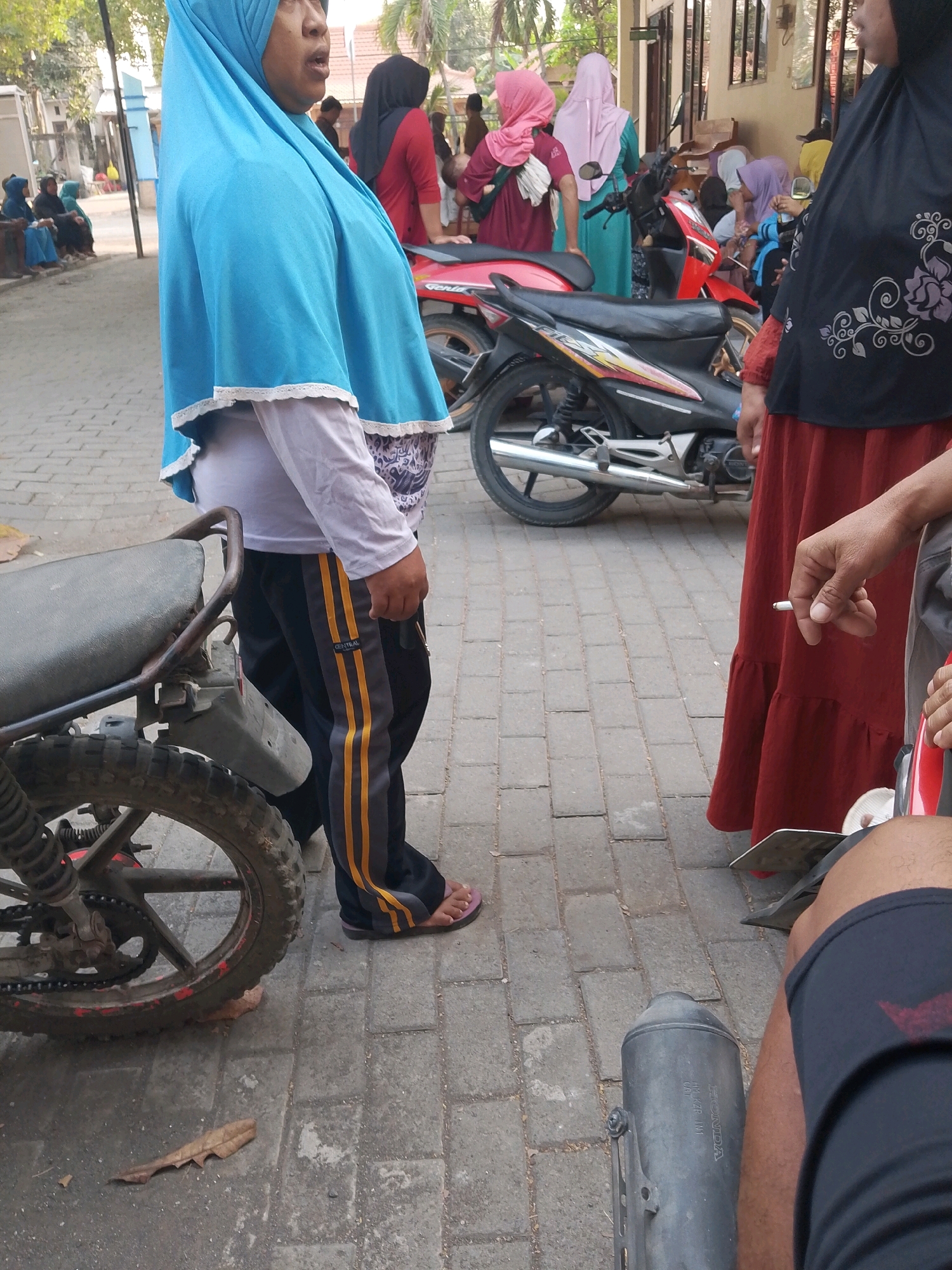 In Wadang, Indonesia, a group of people are gathered near motorcycles on a paved street. The main focus is a woman in a bright blue hijab, wearing a light-colored long-sleeved shirt and striped pants. To her right, another woman in a black hijab and a long red skirt holds a lit cigarette. Several motorcycles are visible, including a black one with red accents and a partially obscured red motorcycle. In the background, other individuals in various colored hijabs and clothing are seen, some seated and others standing. The environment appears to be an outdoor, possibly urban setting, with buildings and trees visible.