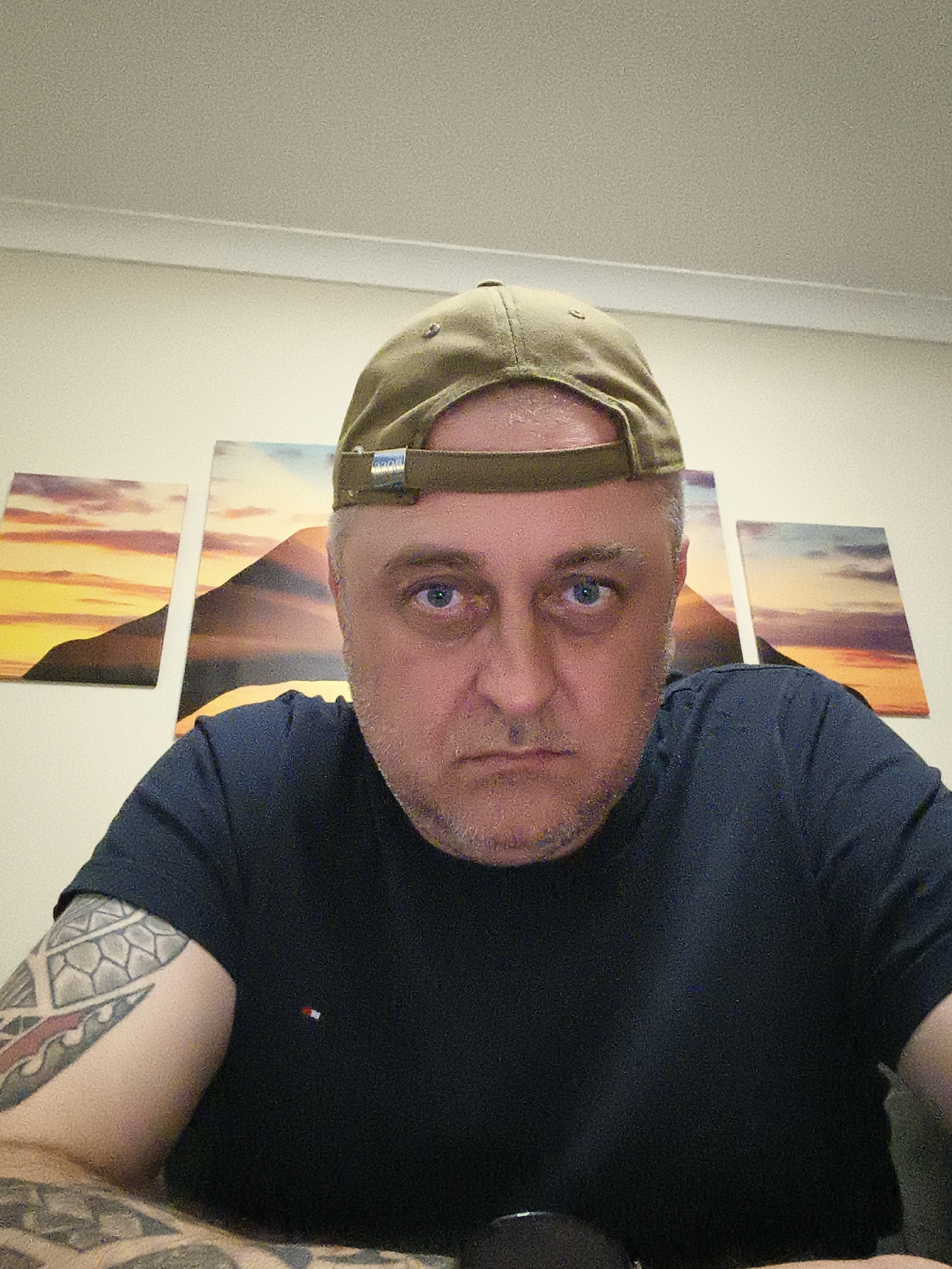 The image presents a close-up, low-angle portrait of a man indoors, likely in Huntingdonshire, United Kingdom. The subject appears middle-aged with fair skin, short grey hair visible beneath his cap, and stubble on his face. He looks directly into the camera with an intense, serious expression in his blue eyes.
The man wears a dark blue, short-sleeved T-shirt with a small, rectangular red, white, and blue striped logo (identifiable as Tommy Hilfiger) on the left chest. His left arm and forearm are adorned with an intricate tattoo featuring geometric and tribal patterns. He is wearing an olive green or khaki baseball cap backward; a metallic clasp with indistinct lettering is visible on the adjustable strap at the back.
The background shows a light-colored wall decorated with three rectangular canvas prints arranged horizontally. These artworks depict a vibrant sunset or sunrise landscape, featuring dark silhouettes of mountains or hills against a sky with warm hues of yellow and orange, blending into cooler purples and blues. Above, a plain, light-colored ceiling with a molding strip is visible. The lighting is even and artificial, illuminating the scene clearly. The overall mood is direct and somewhat introspective.