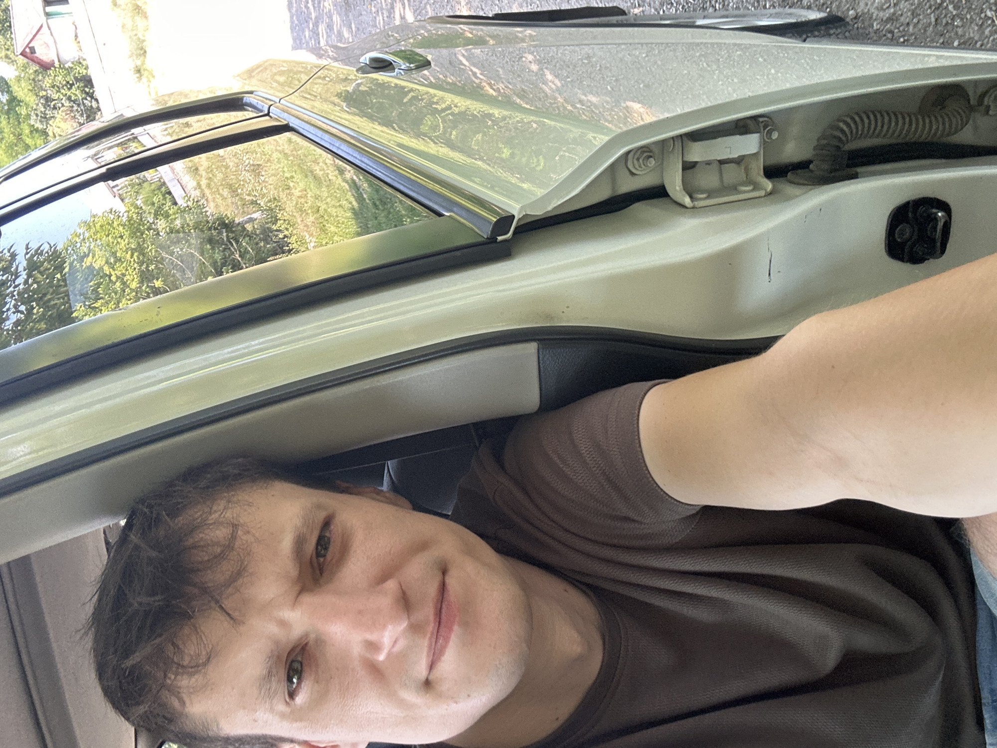A man is seated inside a car with the driver's side door open. He is wearing a brown t-shirt. The car appears to be parked on a gravel surface near a grassy area with trees and houses in the background. The scene is consistent with an outdoor location in Makiv, Ukraine, given the presence of typical residential architecture and vegetation. The lighting suggests it is daytime.