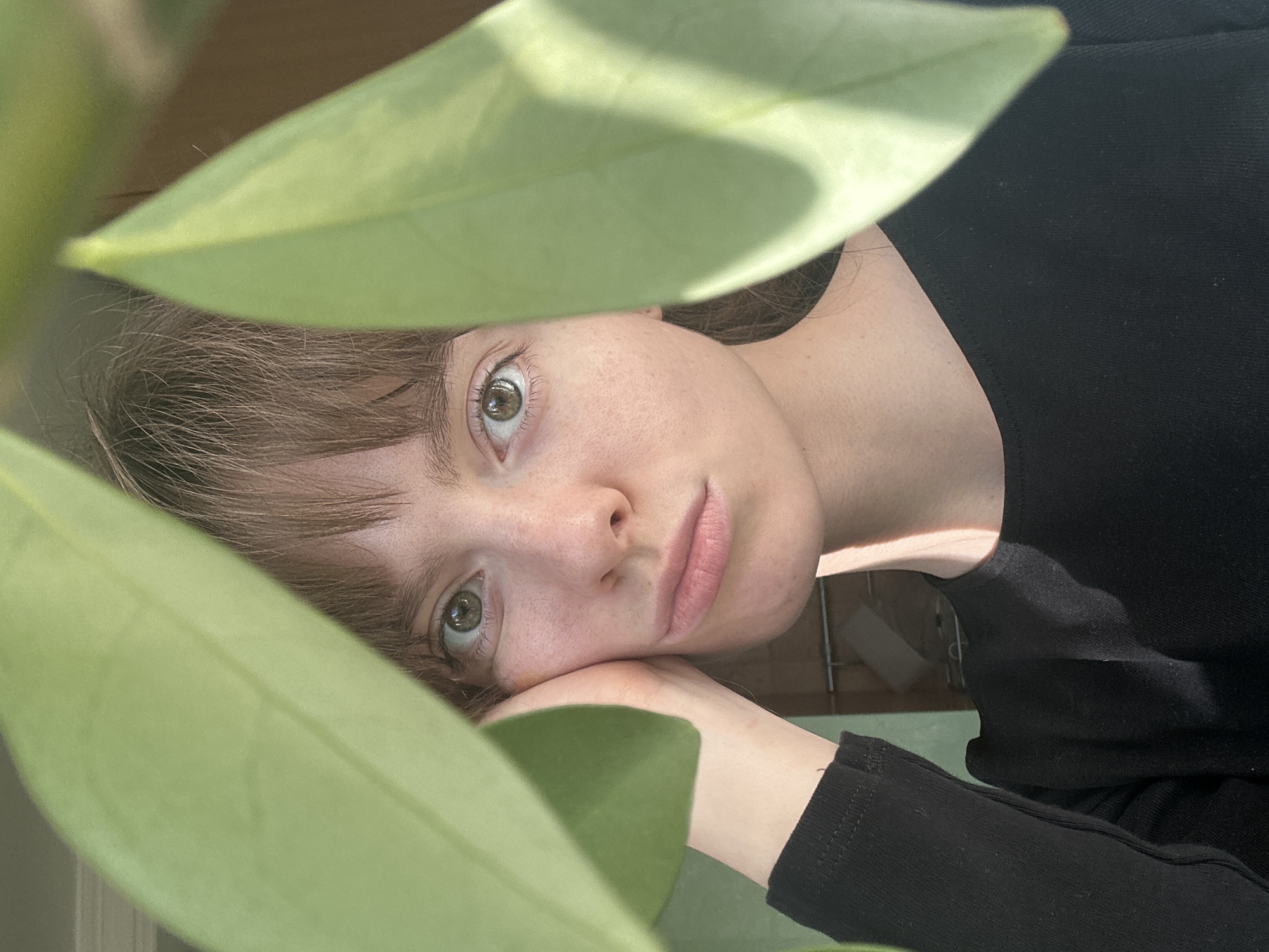 A young person with light skin, brown hair with bangs, and green eyes gazes upward, framed by two large green leaves. Their face rests on their hand. They wear a black, long-sleeved shirt. The background is blurred and suggests a domestic setting with hints of a tiled wall and possibly furniture. The image is likely taken indoors. There is no indication of the location being Moscow, Russia.
