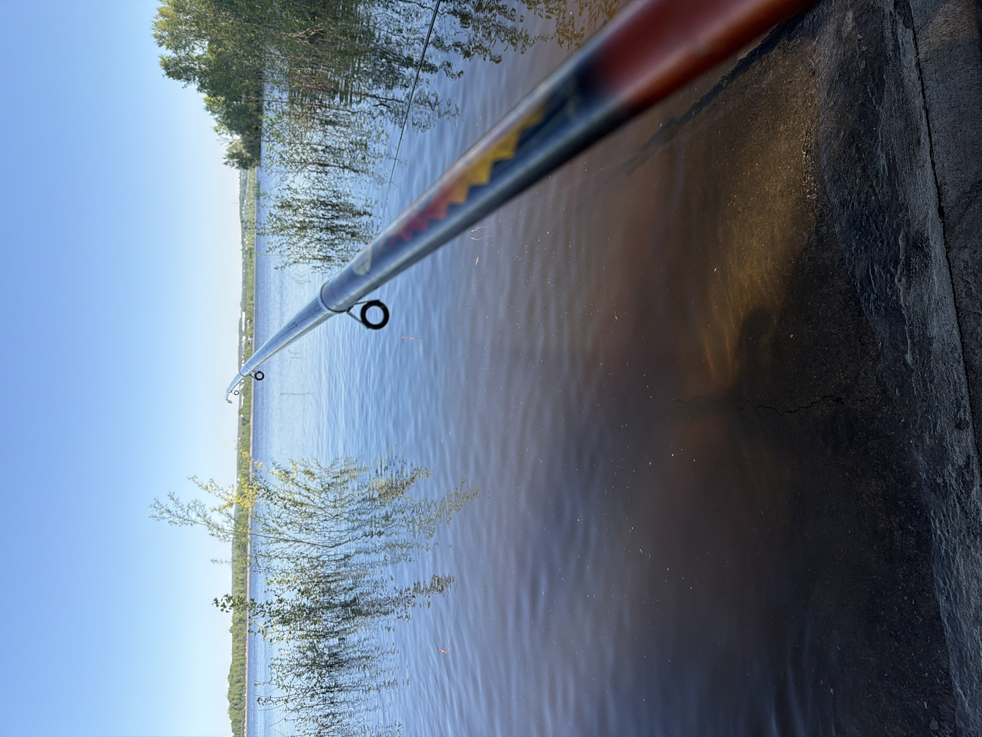 A medium shot depicts a fishing rod extending over a body of water, possibly a river or lake, near Chaikovsky, Russia. The foreground shows a portion of the fishing rod, extending from the bottom right corner diagonally toward the left, into the water. The rod has a colorful decorative band near its handle. The middle ground features the water, reflecting the blue sky and some vegetation along the opposite shoreline. In the background, there is a visible skyline, trees, and a clear sky. On the right, part of a dark-colored surface or ledge is visible. The overall impression is one of calm and natural beauty, suggesting a tranquil fishing scene.