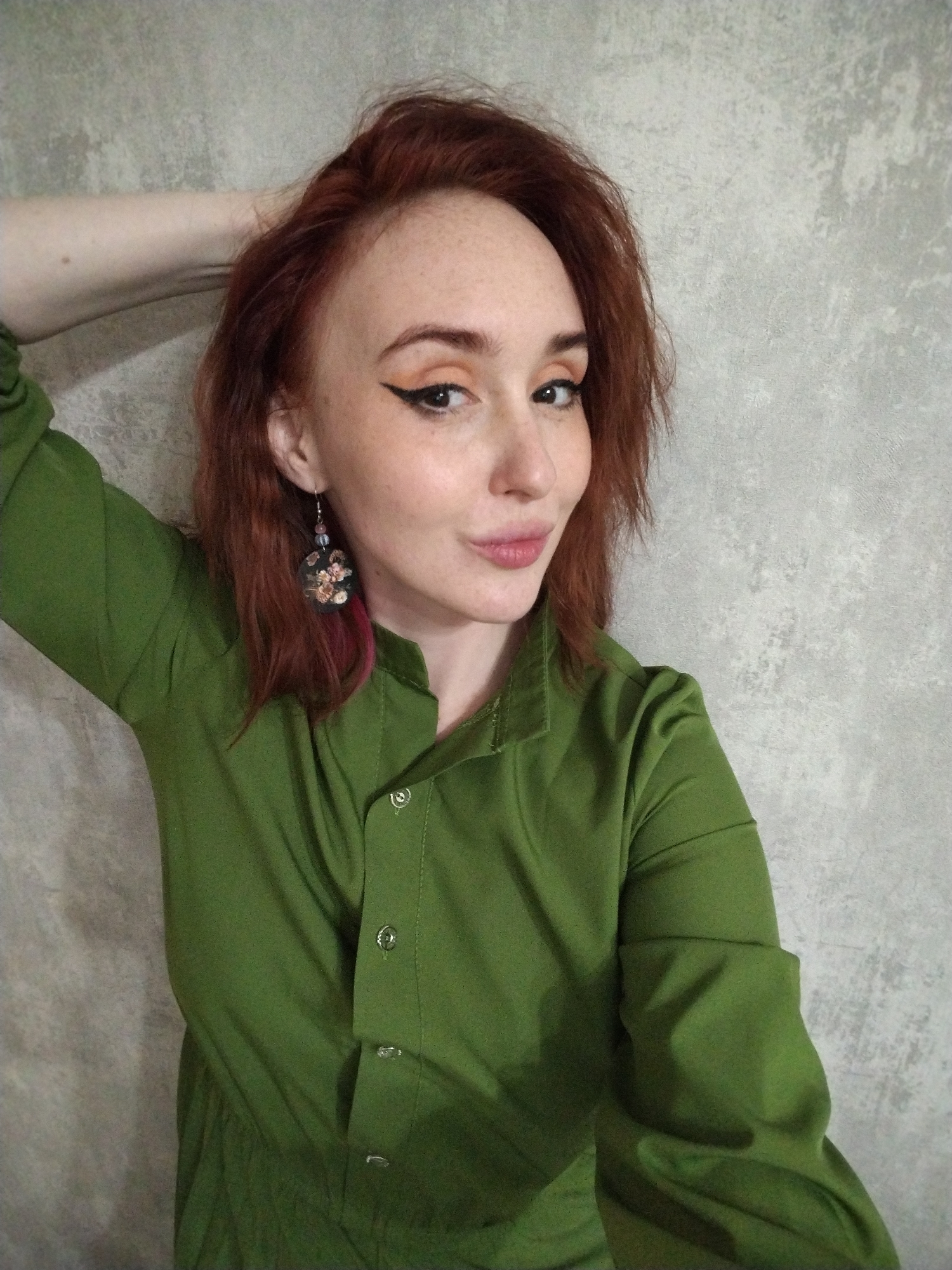 A fair-skinned woman with auburn hair and freckles poses in front of a concrete wall. She wears a long-sleeved olive green shirt dress and dangling earrings. Her makeup includes winged eyeliner. Her left arm is raised above her head, her hand out of frame. The location is unknown and there is no indication of Izhevsk, Russia.