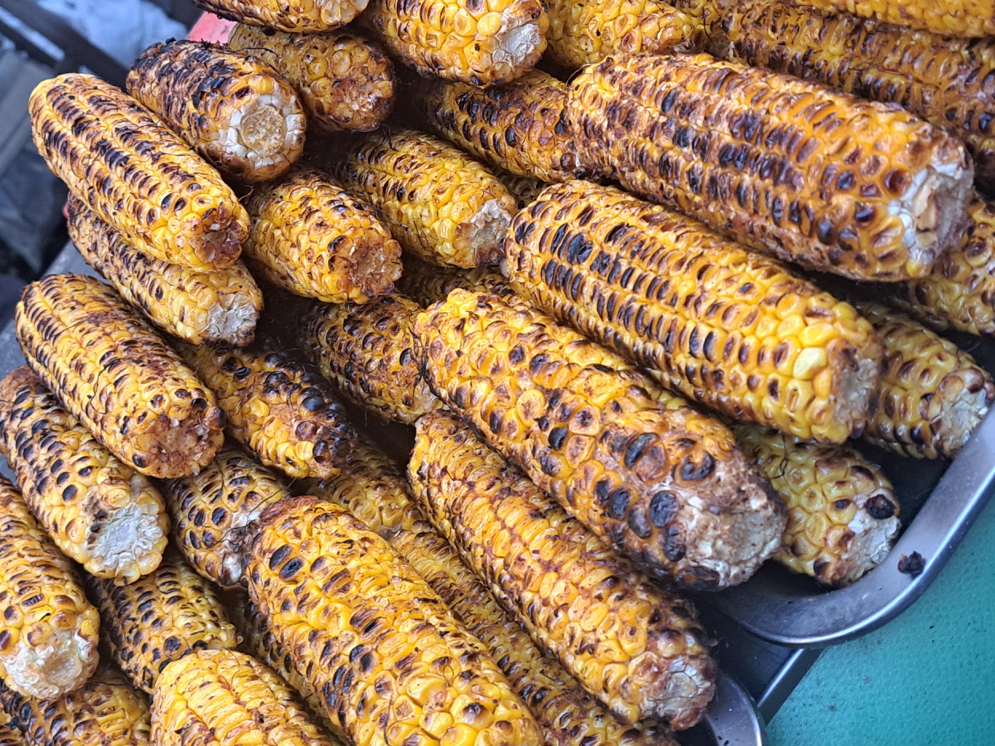 The image displays a close-up, high-angle view of numerous grilled corn cobs, densely piled and filling the majority of the frame. The corn cobs exhibit bright golden yellow kernels with prominent dark brown to black char marks, consistent with grilling. The ends of several cobs are visible, revealing the central white or light brown fibrous core where the kernels terminate. The texture of individual kernels is distinct. The cobs are contained within what appears to be a metallic tray or serving dish, a portion of which is visible along the bottom and left edge. The background is largely obscured by the abundance of corn, with some indistinct darker areas appearing towards the top of the image. A small section of a teal or light green surface is visible to the far left, adjacent to the metallic tray. No people, specific actions, or interactions are visible within the scene. The specific location in the city of Yerevan, Armenia, cannot be determined from the visual information presented in the image.
