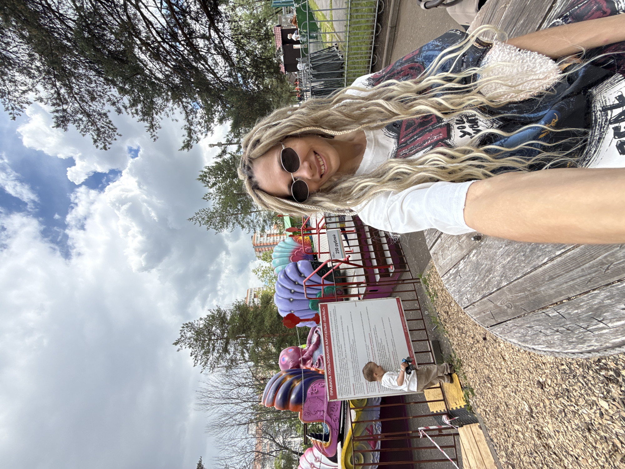 A woman takes a close-up selfie at an amusement park in Rusanovo, Russia, on a partly cloudy day. She has long, blonde dreadlocks and is wearing round sunglasses, a white long-sleeved shirt, and a dark patterned jacket with some white, furry material and partially visible English text, "YOU WAN". She is smiling directly at the camera.
Beside her, a weathered wooden surface, possibly a bench or table, is visible. In the background, an amusement ride featuring colorful, seashell-like structures and an octopus figure can be seen. A young child, dressed in a white t-shirt and tan trousers, stands near a tall white sign next to the rides, holding what appears to be a small toy.
The sign prominently displays "ПРАВИЛА ПОЛЬЗОВАНИЯ КАРУСЕЛЬЮ «ВАЛЬС»" (RULES FOR USING THE "WALTZ" CAROUSEL) at the top, along with a price of "250 рублей" (250 rubles). Detailed rules and regulations are listed below this heading in Russian text. The surrounding area includes green trees, some bare branches, metal fencing, and distant residential buildings. The sky is bright with large white clouds contrasting with patches of blue. The scene depicts a casual, cheerful visit to an outdoor entertainment area.
