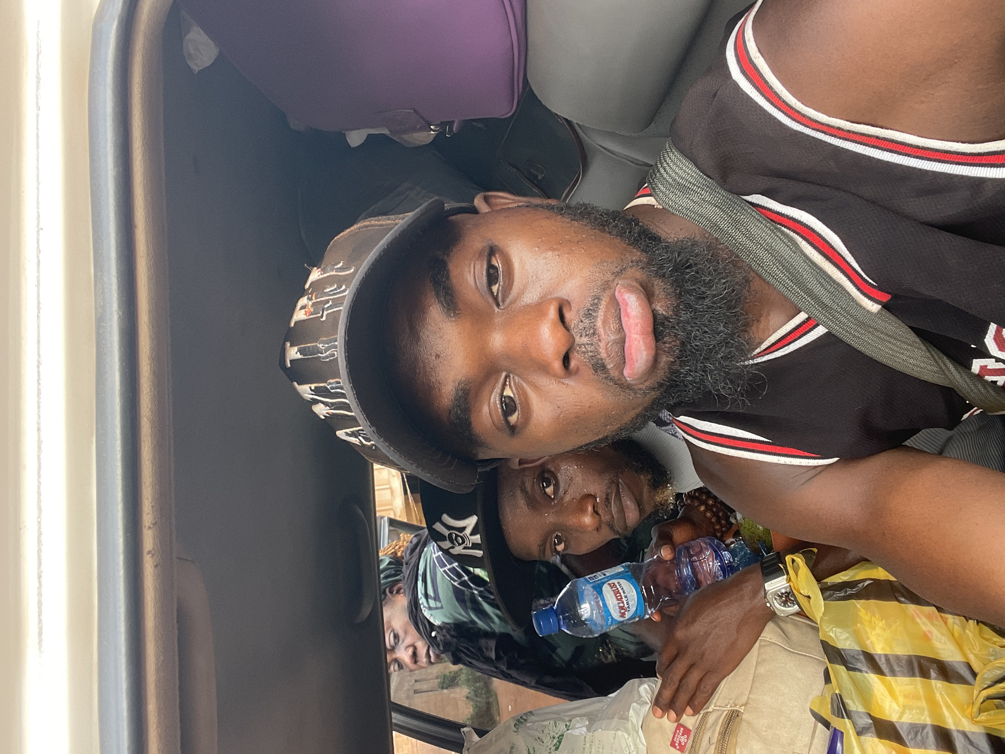 Three men are in a vehicle. The first man, in the foreground, wears a black and red basketball jersey and a hat. A seatbelt is across his chest. He is sticking his tongue out. The second man holds a water bottle. The third man is in the background. A yellow and black striped bag and luggage are also visible. Based on other images from the same source, the location is in Otukpa, Nigeria.
