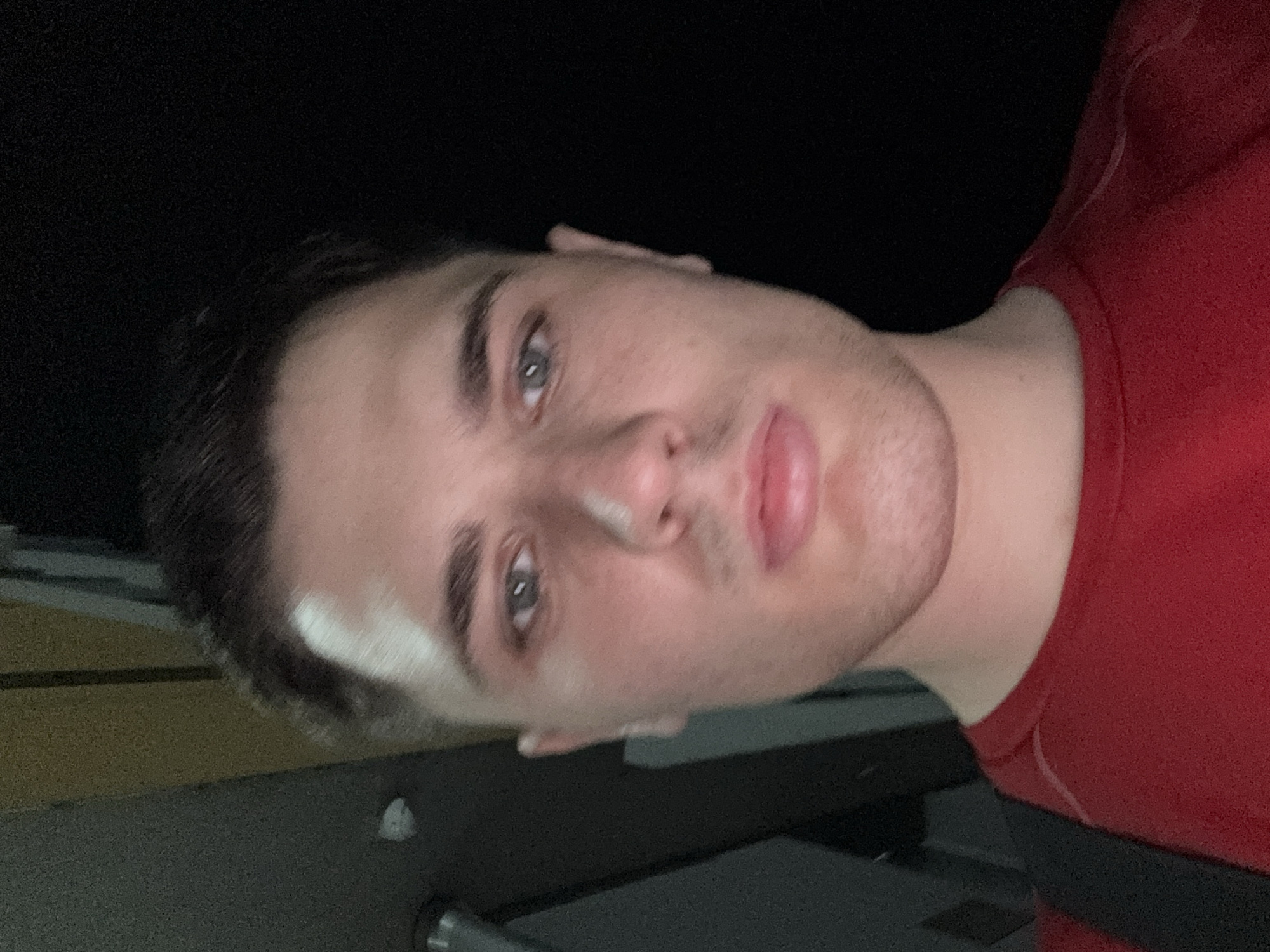 A close-up, high-angle, outdoor selfie shows a young white male wearing a red t-shirt. The subject has short brown hair, fair skin, and blue-grey eyes. The background is dark, with some out-of-focus vertical elements visible on the left side of the frame. No specific location in Stara Zagora, Bulgaria, can be identified from the image.
