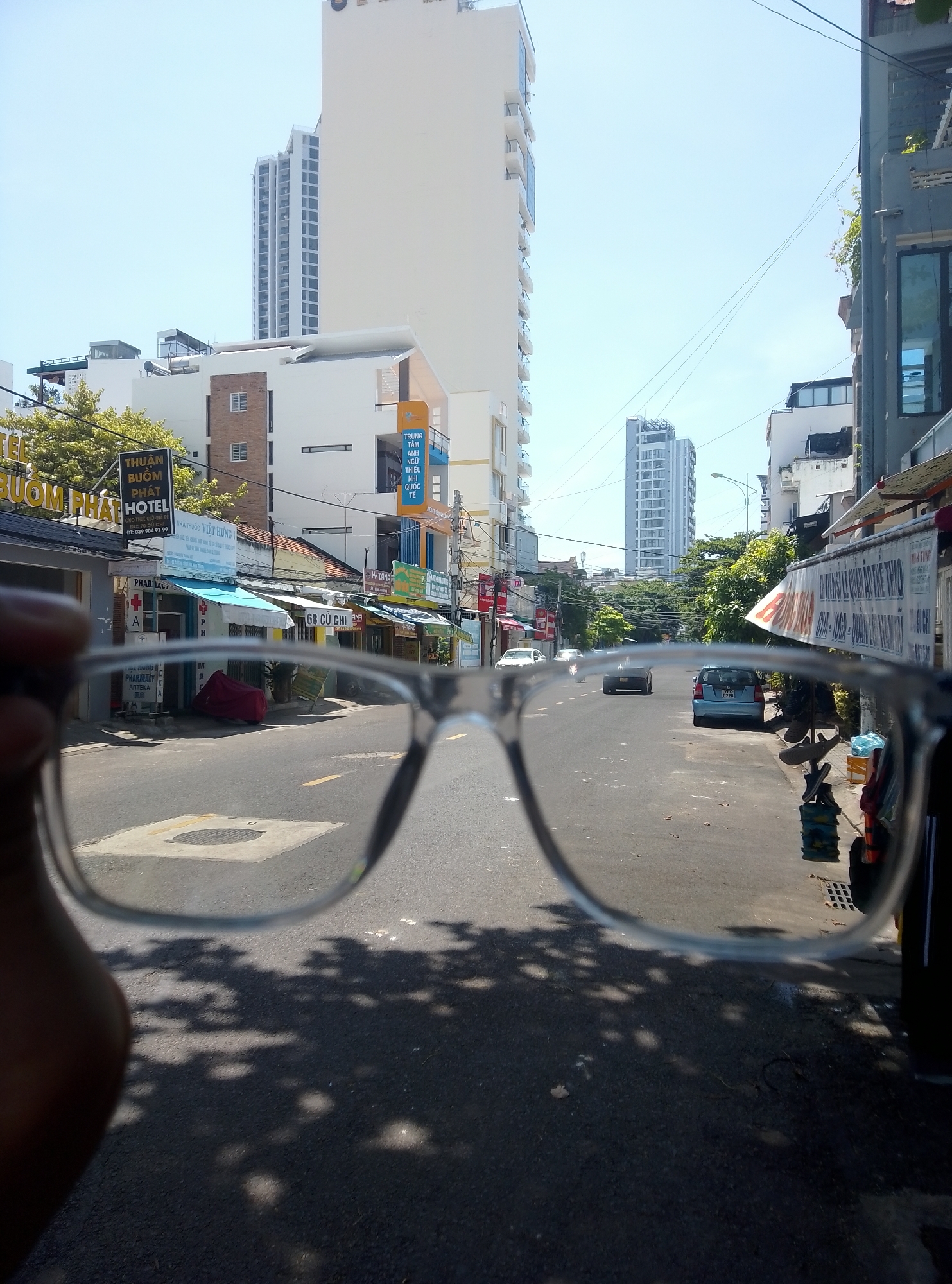 The image captures a sunny street scene in Bac Nha Trang Ward, Vietnam, observed through clear-framed eyeglasses partially held by a human hand in the lower left foreground. The sky is a light, cloudless blue. The asphalt road extends centrally, marked with yellow dashed lines and a square manhole cover in the immediate foreground, exhibiting dappled shadows from unseen trees.
On the left side of the street, a series of buildings includes a prominent white high-rise skyscraper in the background. Closer structures feature commercial storefronts with awnings; identifiable signs read "THUẬN BUÔM PHÁT HOTEL" with a phone number (039 984 97 99), "NHÀ THUỐC VIỆT HÙNG 1" (a pharmacy), and "68 CỦ CHI." A red motorcycle covered by a tarp is parked on the sidewalk.
On the right side, commercial buildings also line the street, including one with an orange and blue sign indicating a "TRUNG TÂM ANH NGỮ THIẾU NHI QUỐC TẾ" (International Children's English Center). A light blue sedan (license plate 43A-012.78) is parked on the right shoulder, facing away from the viewer. Further down the street, two additional cars, one white and one dark, are visible moving away. Numerous overhead power lines crisscross the upper portion of the frame, and a streetlight is visible on the right. Green foliage from trees and bushes is present along both sides of the street.