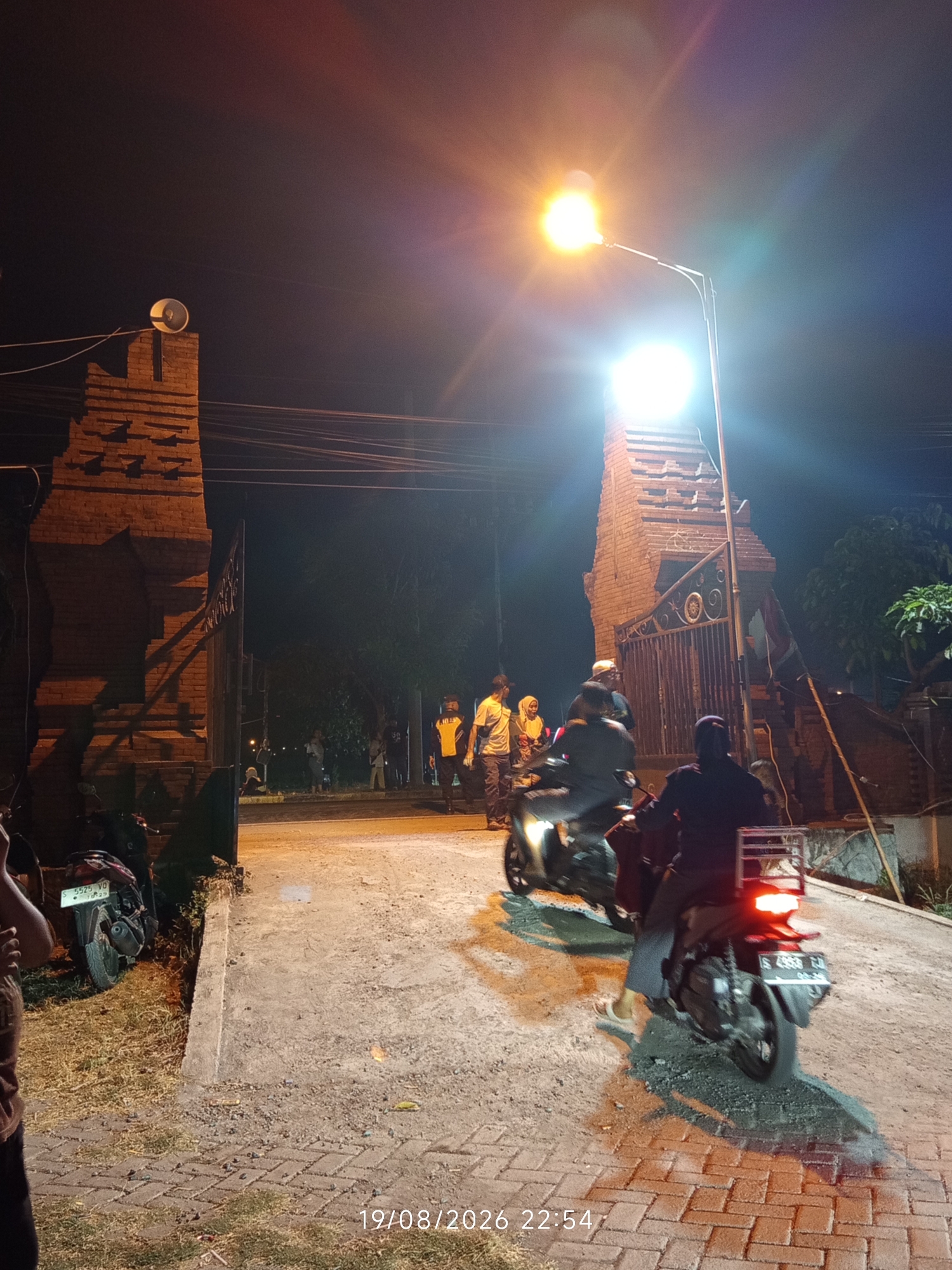 This is a night time, low-angle, medium shot of an entrance gate. The gate is constructed of red brick and features ornate metalwork. A street lamp with a bright light illuminates the scene, casting strong shadows. Two people on a motorcycle are in the foreground, with another motorcycle partially visible to the left. In the background, several other people are gathered near the gate, some standing and some sitting. The ground is a mixture of concrete and brick paving. The timestamp 19/08/2026 22:54 is visible in the lower right corner. The presence of motorcycles and the architecture suggest a location in Indonesia, possibly a residential area or public space.
