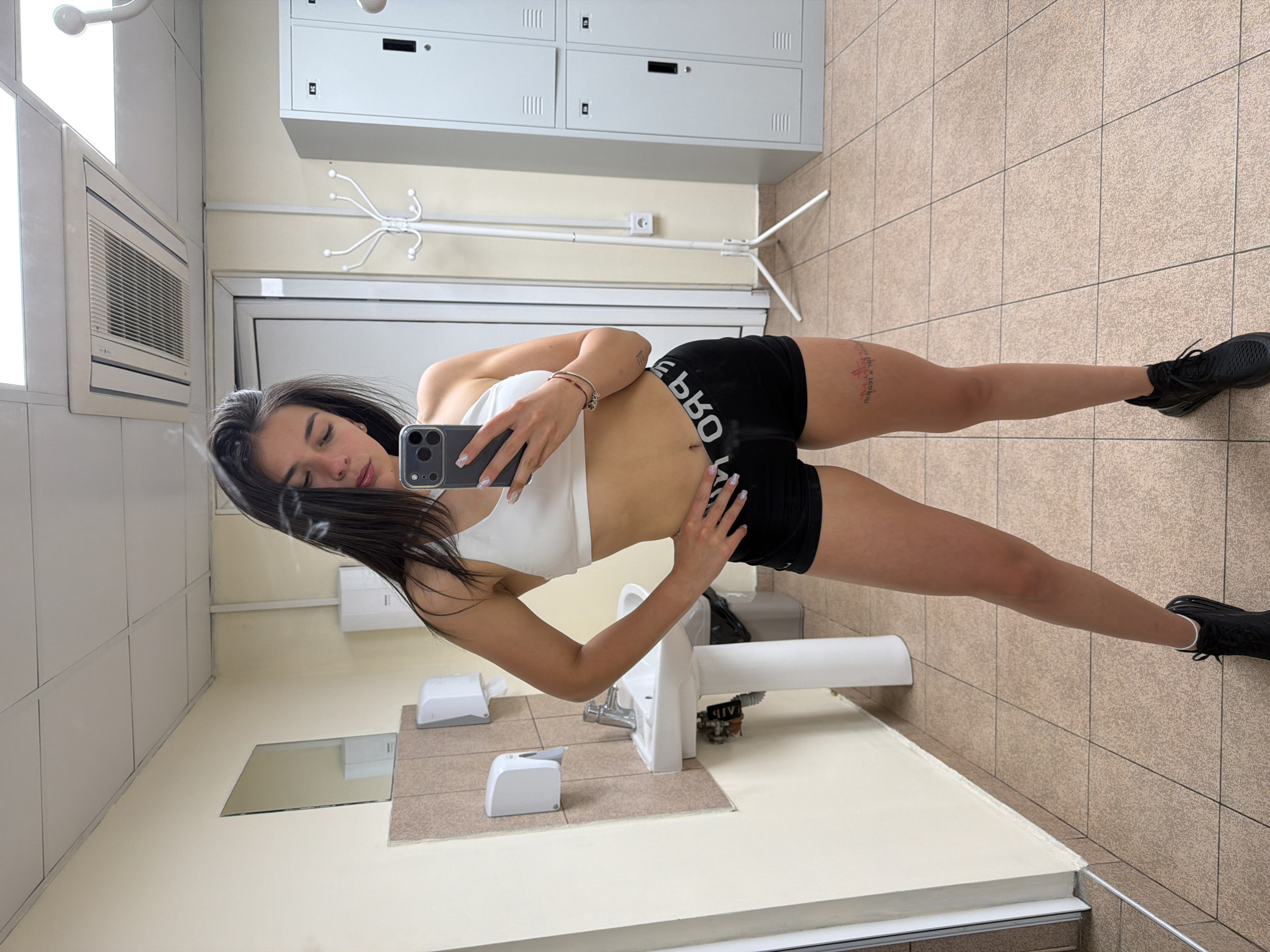 A young woman with long dark hair takes a selfie in what appears to be a locker room or bathroom. She is wearing a white sports bra and black shorts with writing on them and black athletic shoes. She is holding a smartphone. Background elements include lockers, a sink, a towel dispenser, a hand dryer, a coat rack and tiled walls and floor. The location is likely a gym or sports facility. There is no evidence that can place this image in Gabrovo, Bulgaria.