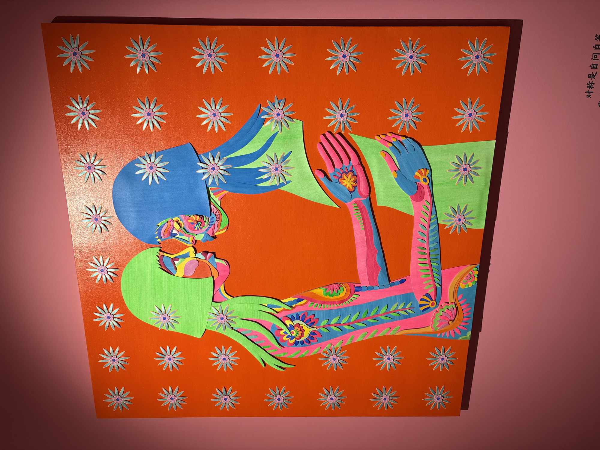 A brightly colored, flat, layered artwork depicts two figures facing each other. The background is orange with scattered, symmetrical flower cutouts. One figure is depicted with blue hair and the other with green. Their faces are detailed with a mix of colors. One figure's arms are raised, showing stylized hands, one of which has a flower design in the palm. The artwork's colors are psychedelic. The image is located in the city of Liangjiang, China.