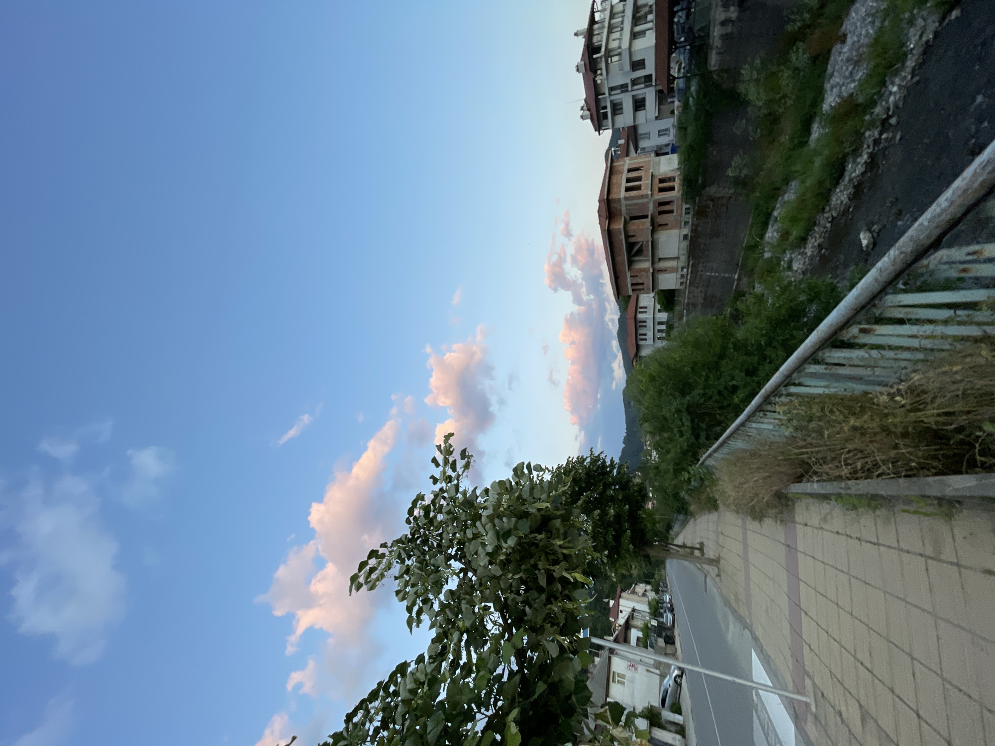The image depicts a street scene in the city of Zlatograd, Bulgaria, during what appears to be late afternoon or early evening. The sky is a clear blue, with scattered, soft pink and white clouds.
On the left side of the frame, a paved sidewalk runs parallel to a road. The sidewalk is made of square tiles, with a subtle pattern of red accents. Trees with lush green foliage line the sidewalk, their branches extending towards the road. Several cars are parked along the side of the road, and a white pedestrian crossing is visible. Residential buildings with white and light-colored facades are situated behind the trees and sidewalk, some featuring red-tiled roofs. One building in the background appears to be under construction, with exposed brick and unfinished windows.
On the right side of the frame, a small, rocky stream bed is visible, partially overgrown with green vegetation. A rusty metal railing runs along the edge of the sidewalk, separating it from the stream area. The background features rolling hills covered in dense green trees. The overall atmosphere is peaceful and serene.