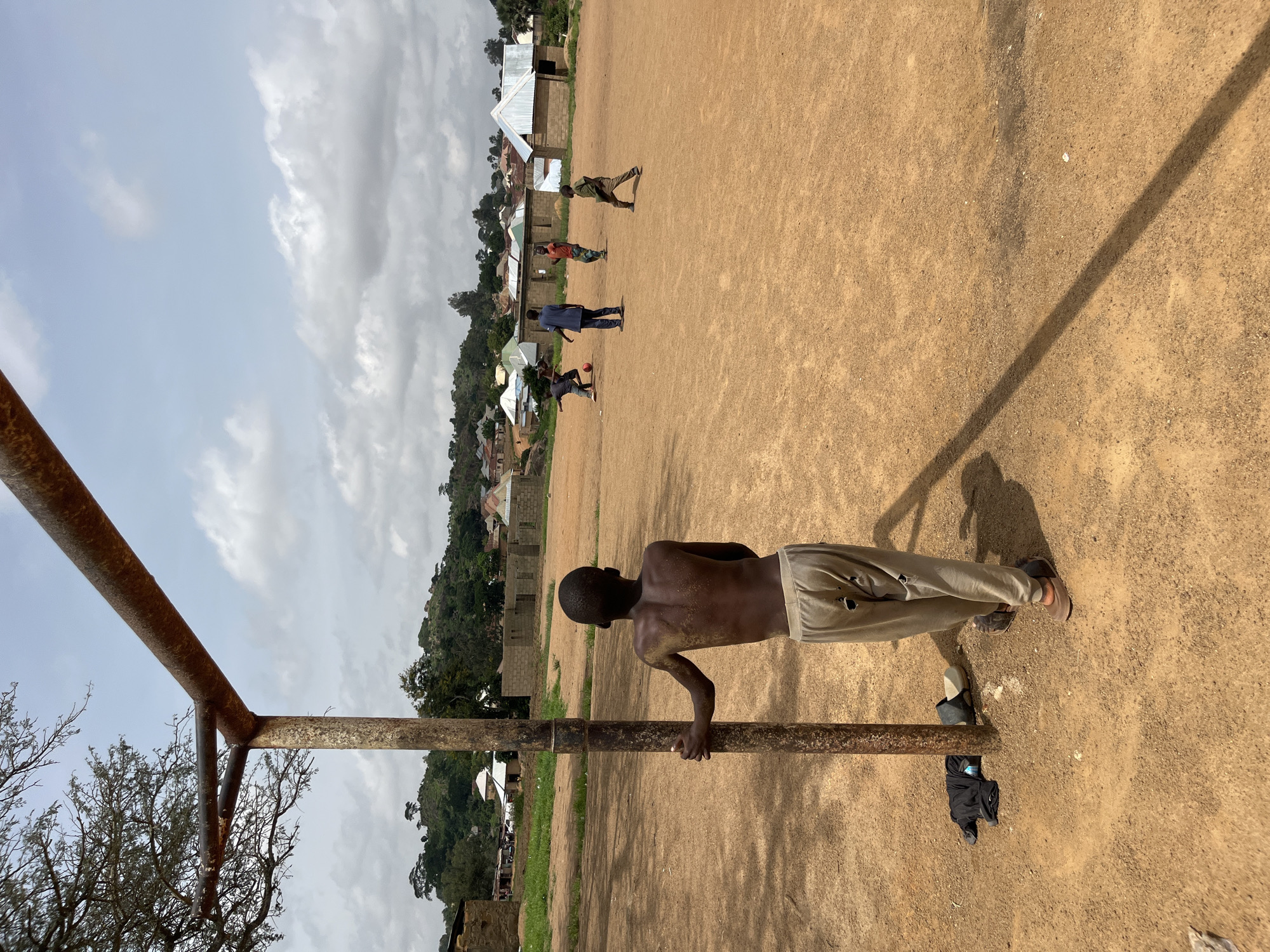In Bukuru, Nigeria, a young boy stands by a rusty goalpost on a dusty, brown soccer field. He is shirtless, with his back to the camera, wearing light-colored, torn pants and brown sandals. His skin appears to have some discoloration or dirt on his upper back. To his left, a pair of dark sandals and a dark t-shirt lie on the ground.
In the background, several other boys are playing soccer. One boy is kicking a red soccer ball, while others run and position themselves on the field. A man in a blue shirt and dark pants stands watching the game. The environment is rural, with several mud-brick buildings and some houses with corrugated metal roofs visible in the distance. Trees and vegetation dot the landscape, and a gentle hill rises behind the buildings. The sky is partly cloudy with some white clouds scattered across a light blue expanse. The overall scene depicts a casual soccer game in a village setting.
