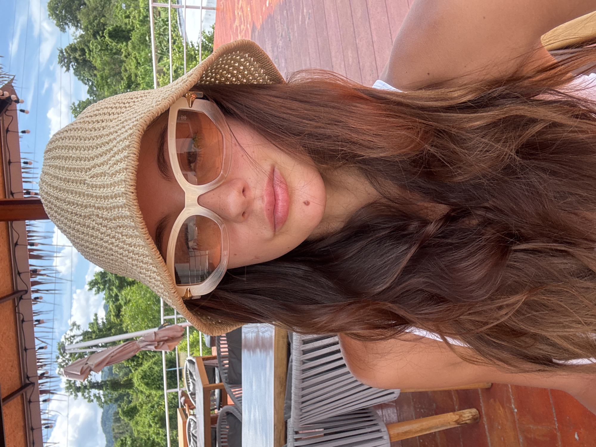 A woman with long brown hair is wearing a beige straw hat and off-white framed sunglasses. She has a small mole on her left cheek. The setting appears to be an outdoor patio or deck. Chairs and a table are visible. In the background, there is lush green vegetation and what appears to be a hill or mountain. The location is likely Kathu, Thailand.