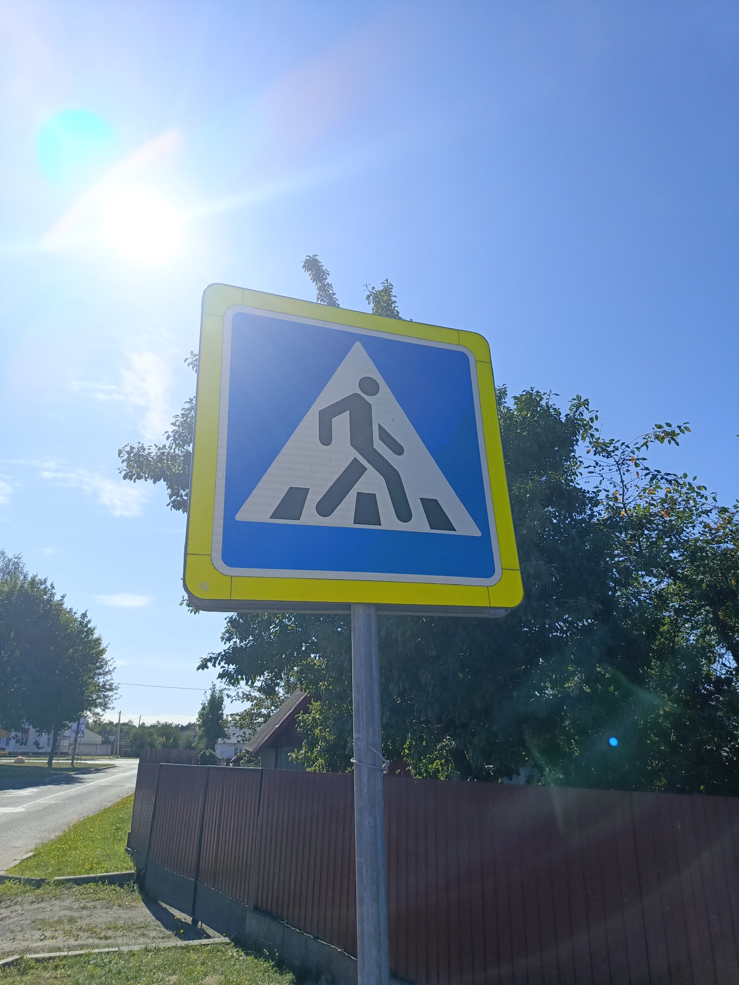 A pedestrian crossing sign is shown against a bright blue sky with sun glare. The sign is a blue triangle with a white border and a yellow outer border. Inside the triangle, a black silhouette of a person is walking across white stripes representing a crosswalk. The sign is mounted on a metal pole. In the background, there are green trees, a brown wooden fence, a road, and some buildings. The scene appears to be in a suburban or rural area. The location is identified as Ivatsevichy, Belarus.