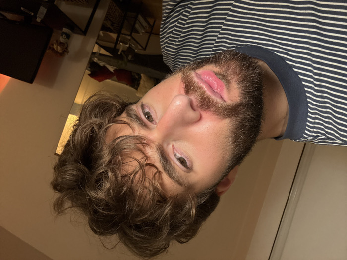 A close-up, tilted portrait-style image captures a male individual, appearing to be in his 20s or 30s, positioned in an indoor setting. He has medium skin tone, light brown/hazel eyes, a thick dark brown beard, and thick, curly dark brown hair. He wears a navy blue and white striped t-shirt with a solid navy collar. His gaze is directed towards the camera with a neutral expression. The lighting is warm and artificial, illuminating the subject and the immediate background.
To the right of the subject, a tall, narrow mirror reflects a portion of the room, revealing a dark sofa adorned with red cushions, situated behind a black coffee table. Further right, a dark television screen rests on a black stand. A warm orange glow, consistent with an ambient light source, is present at the extreme right edge of the frame. The walls are a light, neutral color. The environment is consistent with a private residential interior. No specific visual cues within the image indicate the location is Paris, France.