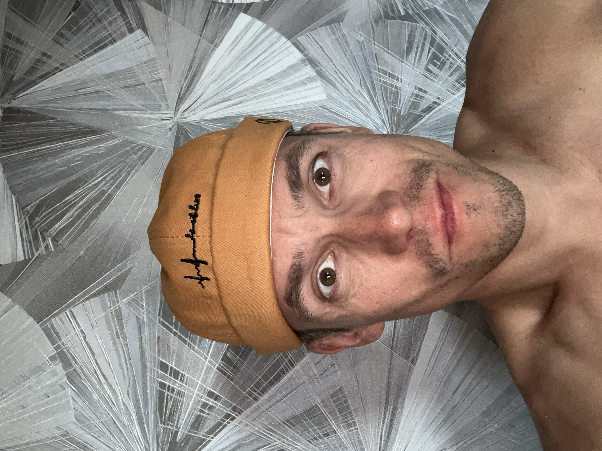 The image, rotated 90 degrees clockwise, presents a close-up portrait of a man from the chest up, set against a distinct patterned wall in Chaikovsky, Russia.
The man, appearing to be in his 20s or 30s, has fair skin and is shirtless, revealing his shoulders and upper chest. He has short stubble or a closely trimmed beard on his chin and upper lip. His most striking features are his wide, intense brown eyes that look directly at the viewer, conveying an alert or surprised expression. His lips are slightly parted and appear reddish. He is wearing an orange-brown or ochre-colored beanie, typical of a dockworker cap, with its brim folded up.
On the front of the beanie, there is black embroidered text in a stylized cursive font, which appears to read "Brooklyn."
The background is a wall covered with a light gray and white patterned wallpaper. The pattern is abstract and geometric, characterized by lines radiating outwards from central points, creating a crystalline or fan-like effect. The lighting appears even, suggesting an indoor setting. The man's direct gaze into the camera suggests he is taking a selfie or posing for a photograph.