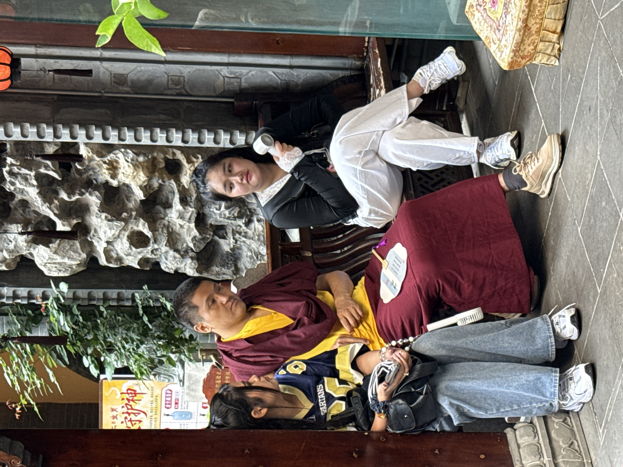 Three people are seated in what appears to be a covered outdoor or semi-indoor area, possibly a courtyard or cloister, with stone walls and traditional-style wooden benches. The image is rotated 90 degrees clockwise.
In the center, a man wearing maroon and yellow monastic robes, consistent with those of a Buddhist monk or lama, sits with his arms crossed, looking towards the left. On his lap rests a paper fan and white prayer beads. To his left, a young woman is seated, wearing a blue and yellow sports jersey with "SPARTANS" visible on it, light blue wide-leg jeans, and white sneakers. She is looking downwards, holding a black shoulder bag and possibly her phone. To the man's right, slightly behind and higher up, another young woman is seated. She wears a black long-sleeved shirt with white lace cuffs and collar, white pants, and white sneakers. She holds a small white portable fan and looks towards the viewer.
The setting includes decorative stone carvings on the wall, lush green foliage hanging from above, and a tiled floor. A yellow sign with Chinese characters is partially visible in the background, displaying "守护神" (Shǒuhùshén), meaning "Guardian God" or "Protector God." The people appear relaxed, suggesting they are resting or taking a break. The presence of a portable fan hints at warm weather.