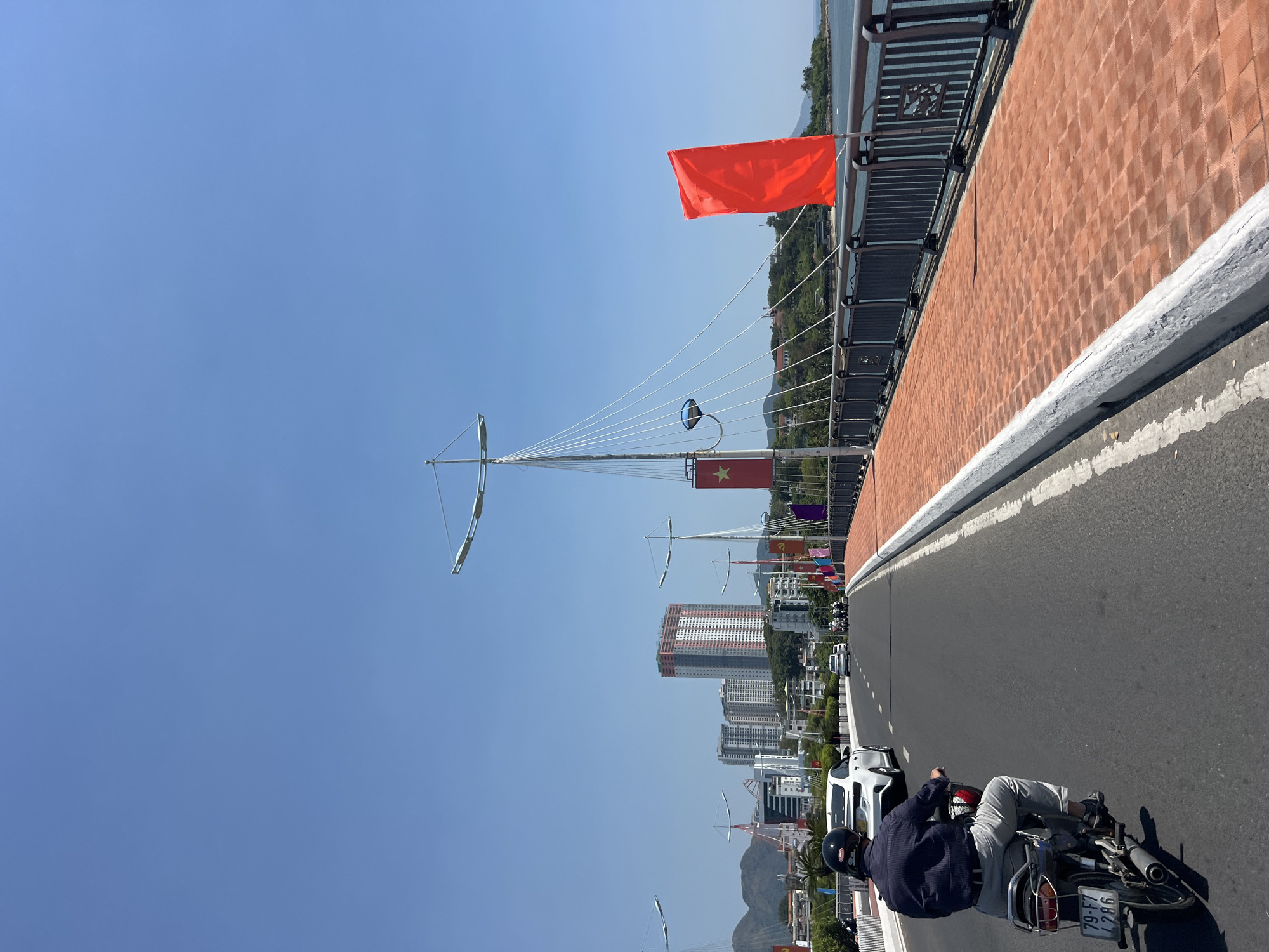 The photograph, taken in Nha Trang, Vietnam, shows a person riding a motorcycle on a bridge. The bridge has red brick side railings, a white concrete border, and a dark asphalt road surface. Several red and communist Vietnamese flags are attached to the bridge's railing and cables. In the background, the cityscape of Nha Trang includes modern high-rise buildings and distant mountains. The sky is clear blue. The motorcycle rider is wearing a dark jacket, light pants, and a helmet. The motorcycle's license plate is partially visible.
