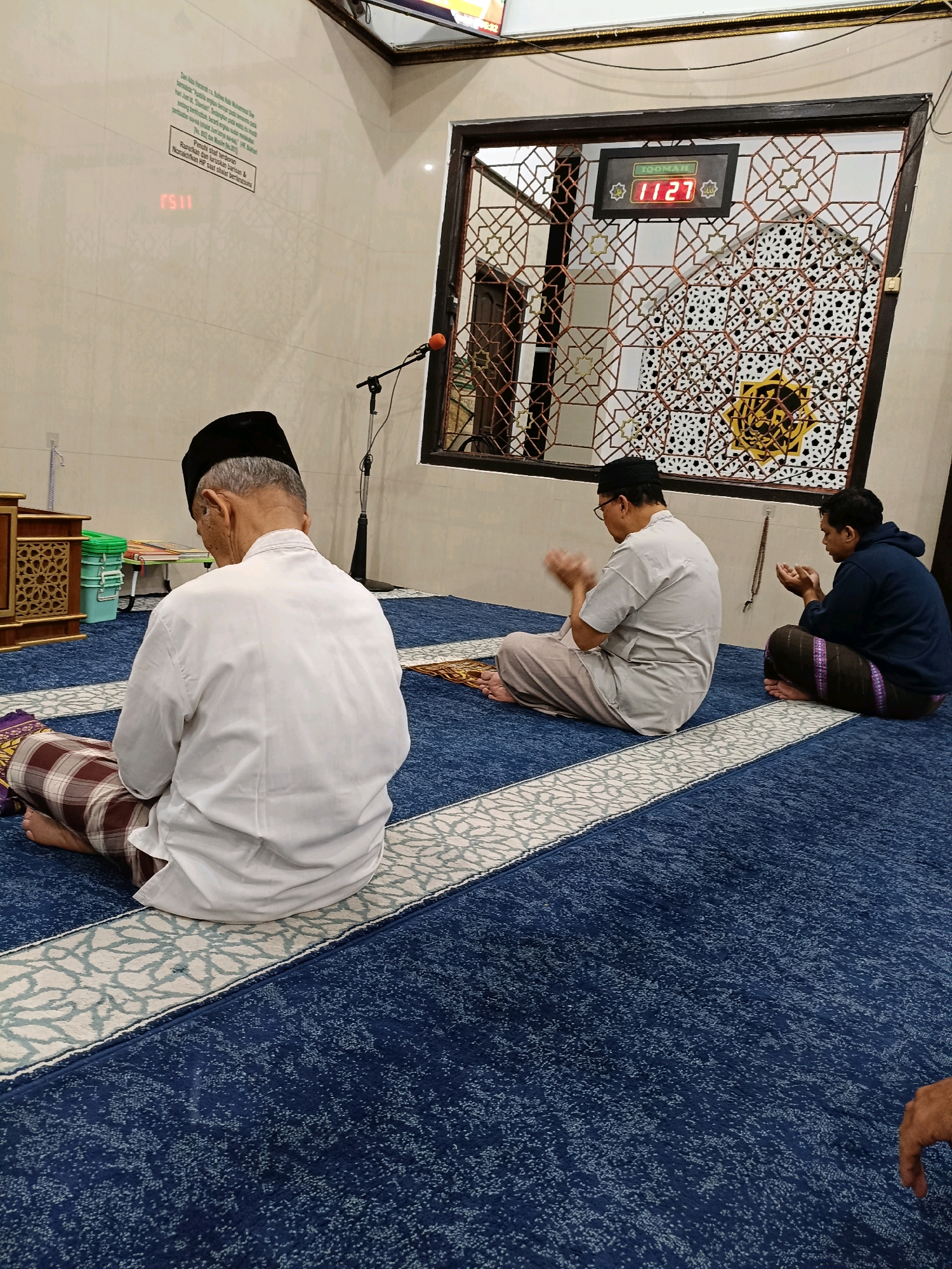 Three Muslim men are praying on a blue carpet in a room that appears to be a mosque. The man in the foreground, wearing a white shirt and black cap, is facing away from the camera. Two other men are seated in front of him, also in prayer. The man in the middle is wearing glasses and a light-colored shirt, with his hands raised. The man on the right is wearing a dark blue hoodie and has his hands cupped. Above them, a digital clock displays "11:27". The walls are tiled, and a decorative metal screen is visible in the background. The overall scene suggests a place of worship, possibly in Maguwoharjo, Indonesia, given the context.