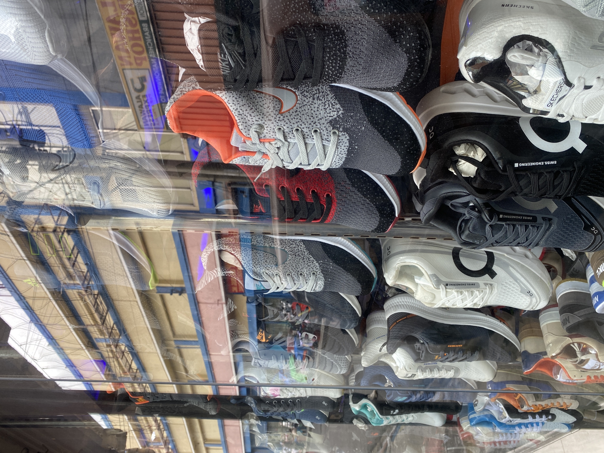 The image shows a display window of a shoe store in Davao City, Philippines. The window is filled with various athletic shoes, mostly sneakers, arranged in vertical rows. The shoes are predominantly in shades of gray, black, white, and red, with some featuring bright accents like orange and blue. Brands like Nike and Skechers are visible on some of the shoes. The display is organized, with shoes placed side-by-side and some stacked. Reflections of buildings and possibly other shops are visible on the glass of the display window, suggesting it's located in a commercial area. The lighting appears to be natural daylight, with some internal store lighting also contributing. No people are clearly visible interacting with the display, indicating the photo was taken from outside the store. The background visible through the window shows building facades with windows and signage, consistent with an urban environment.