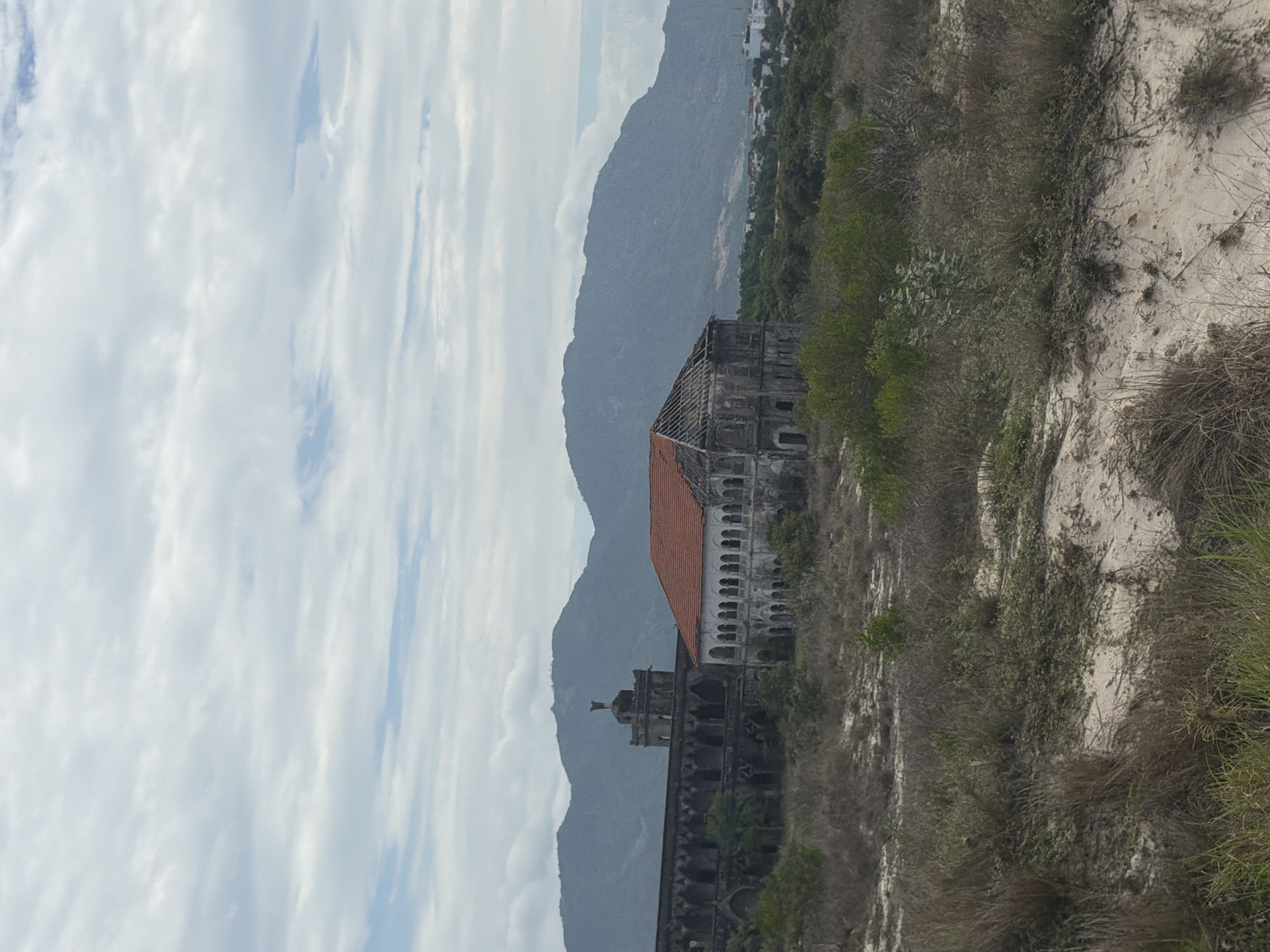 An expansive landscape shot captures a weathered, possibly abandoned colonial-era building set against a backdrop of mountains and a cloudy sky in Bac Cam Ranh Ward, Vietnam.
In the mid-ground, the main subject is a large, multi-story stone or concrete structure with distinct architectural features. One prominent section features a sloping red-tiled roof and multiple arched windows or openings on its facades, suggesting a European influence. To the left of this section, another part of the building, possibly a tower, rises higher, capped with a small spire or statue. The entire building shows signs of age and disrepair, with faded surfaces and missing elements.
Behind the building, a long range of dark, hazy mountains stretches across the horizon, appearing rugged and imposing. Further in the distance, to the right and nestled among the mountains, a cluster of smaller, modern buildings indicates a distant town or settlement.
The sky above is largely covered with white and grey clouds, though patches of clear blue sky are visible, suggesting it is daytime. The clouds appear somewhat streaky.
On the right side of the foreground, a sandy or light-colored scrubland rises, covered with sparse, dry and green vegetation. Various bushes, some vibrant green and others brown and withered, along with a few cactus-like plants, dot the arid slope.
No people or specific activities are visible, and there is no discernible text in the image. The scene conveys a sense of history and nature's reclaiming presence.
