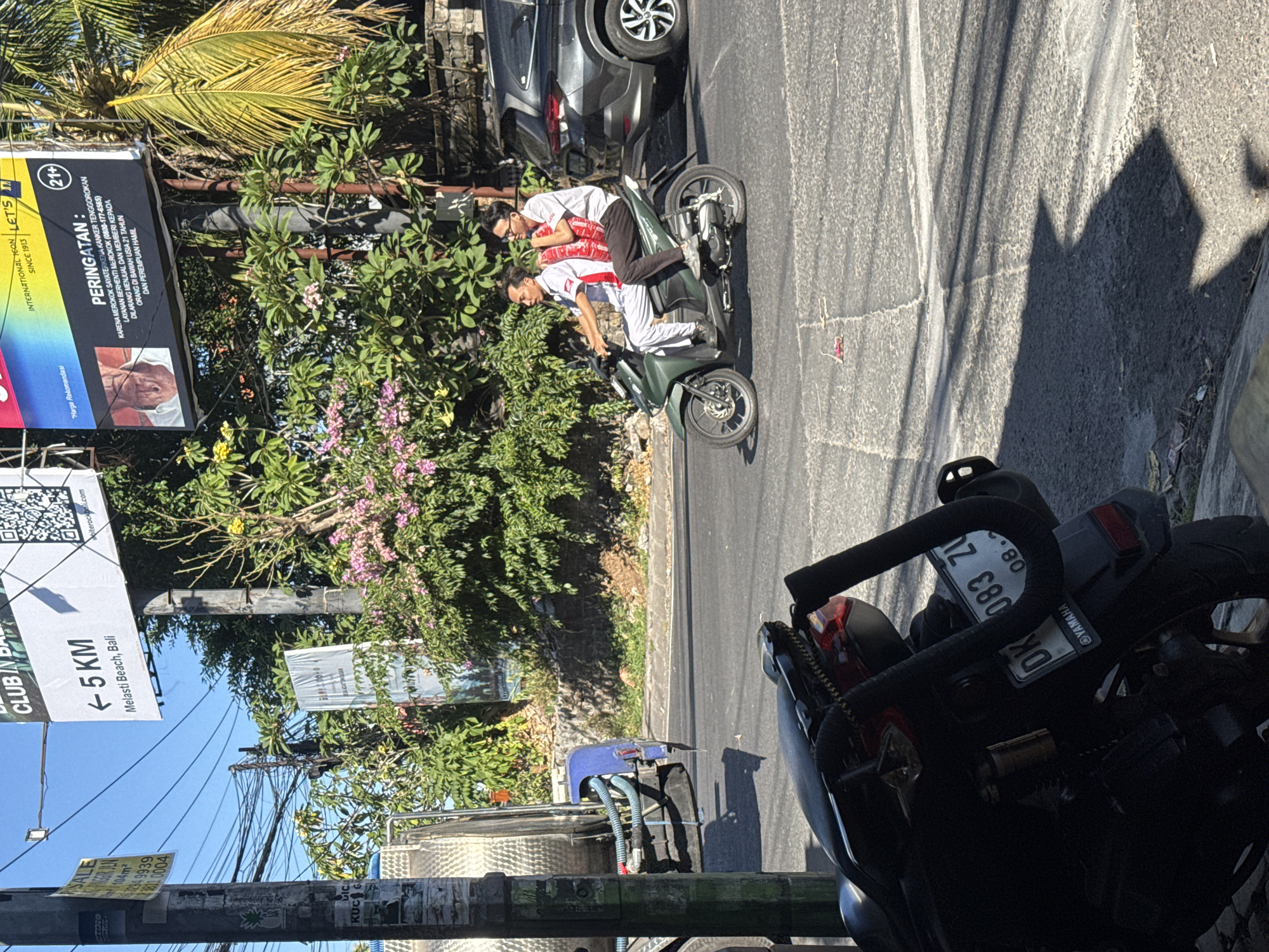 The image depicts a street scene in Kuta Selatan, Indonesia, on a sunny day. In the foreground, a dark-colored motorcycle is parked, partially obscuring the view of the street. Two men are riding a green scooter on the road. The driver, wearing glasses and a white shirt, is looking forward, while the passenger, in a white shirt with red accents and dark pants, is looking to his left. The passenger is carrying two large red plastic bags. Behind the scooter, a dark-colored SUV is visible. In the background, there are several poles with signs and wires, and lush green foliage with pink flowering bushes. A large billboard with Indonesian text and an advertisement for a product with a "21+" rating is also visible. The road surface appears to be asphalt with painted white lines.