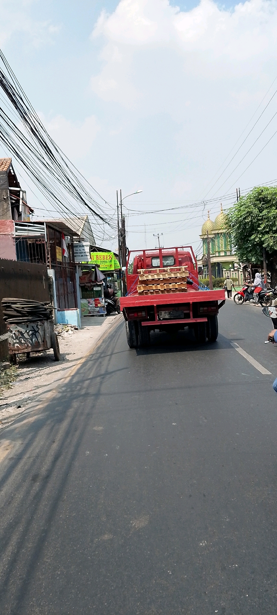 A red flatbed truck, loaded with stacks of wood, drives down a two-lane asphalt road in Bekasi, Indonesia. The truck is the primary subject, viewed from behind and slightly to the right. In the background, on the left side of the road, is a building with a red roof and an Indonesian flag hanging on its facade. A sign with Indonesian text, "GET CLINTON" and "BEBEK," is visible on the building. Further down the road on the right, a mosque with golden domes stands out against the sky. Several motorcycles are present on the road; one is parked on the left, and two others are in motion on the right, one carrying two people. The sky is a bright blue with scattered clouds. The environment appears to be a street in a bustling urban or semi-urban area, with overhead electrical wires crisscrossing the scene.