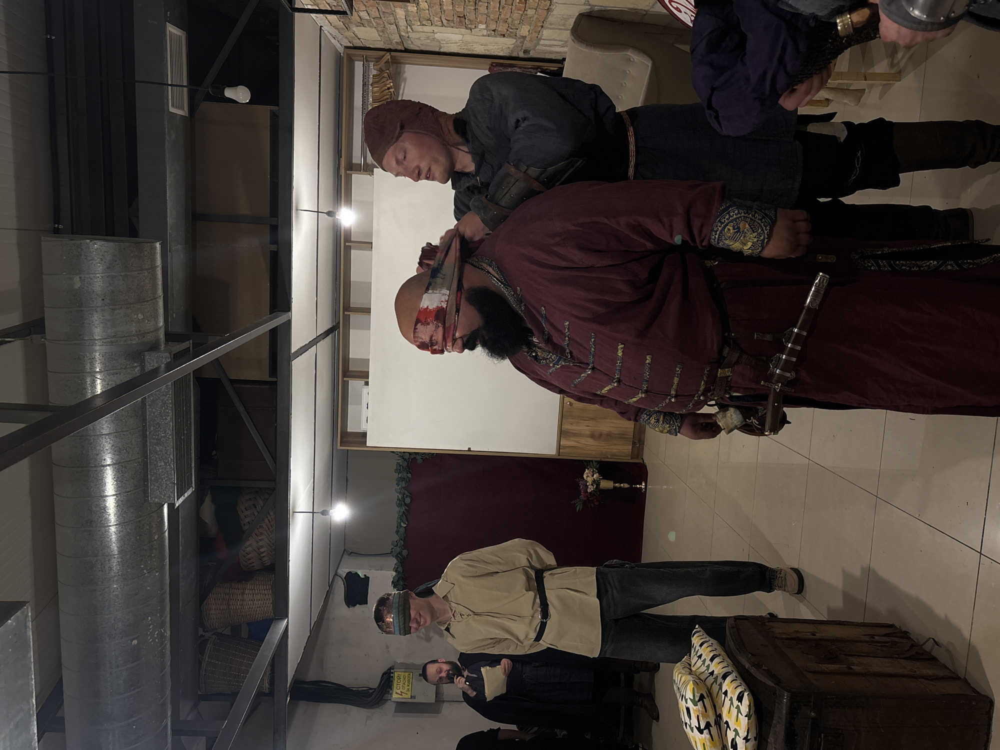 The image shows an indoor scene with several individuals dressed in what appears to be historical or theatrical costumes. One man, wearing a red and gold robe-like garment, has a blindfold being secured around his head by another man dressed in darker medieval-style clothing. A sword is visible near the robed man. In the background, another person in similar attire stands near a chest. The location is difficult to ascertain definitively but the presence of tiled floors and the decor suggests it could be a themed event or reenactment taking place within a building, possibly in a city like Varna, Bulgaria. Further specific forensic details are not discernible from the image alone.