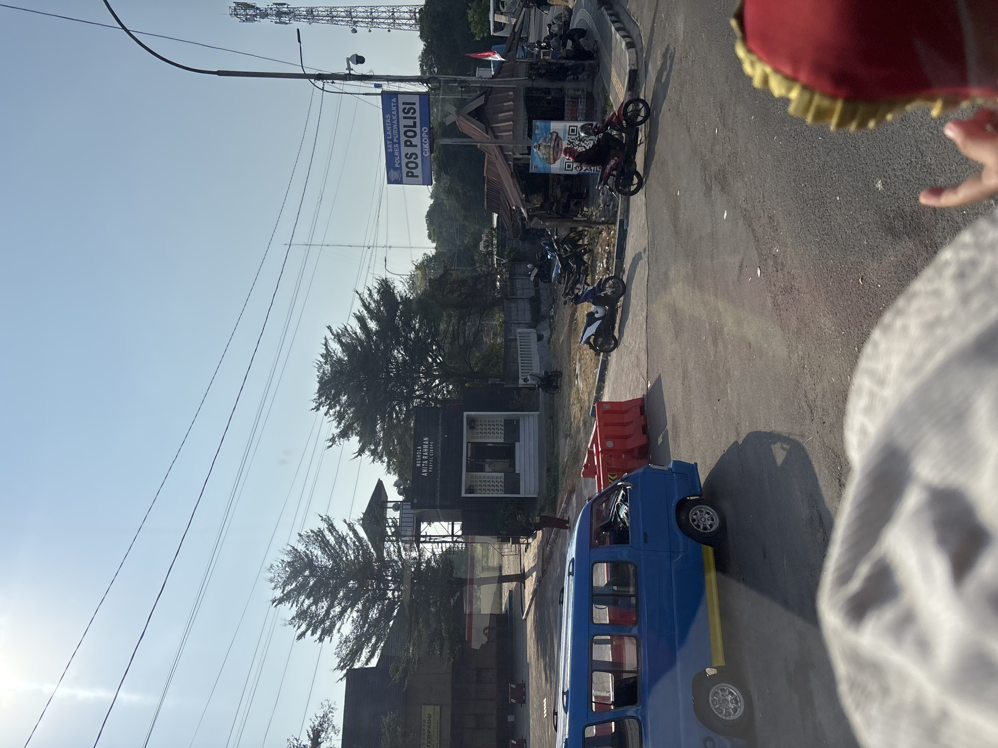 In Cikampek, Indonesia, a clear, sunny day is depicted. In the foreground, the blurry image of a person wearing a white headscarf and a red cap with gold trim is visible, partially obstructing the view. A blue minibus with yellow trim is parked on the right side of the road. Several red plastic barriers are placed on the road.
Behind the minibus, a small building with the text "MUSHOLLA ANITA RAHMAN POSPOL CIKOPO" is visible. To the right of this building, a larger sign reads "SAT LANTAS POLRES PURWAKARTA POS POLISI CIKOPO". Adjacent to the "POS POLISI" sign is a small food stall with a roof made of corrugated metal, and several motorcycles are parked nearby. A man on a red motorcycle is seen riding away from the stall, while other motorcycles are parked. A cell tower is visible in the background on the right. The sky is clear and blue, with overhead power lines crisscrossing the scene.