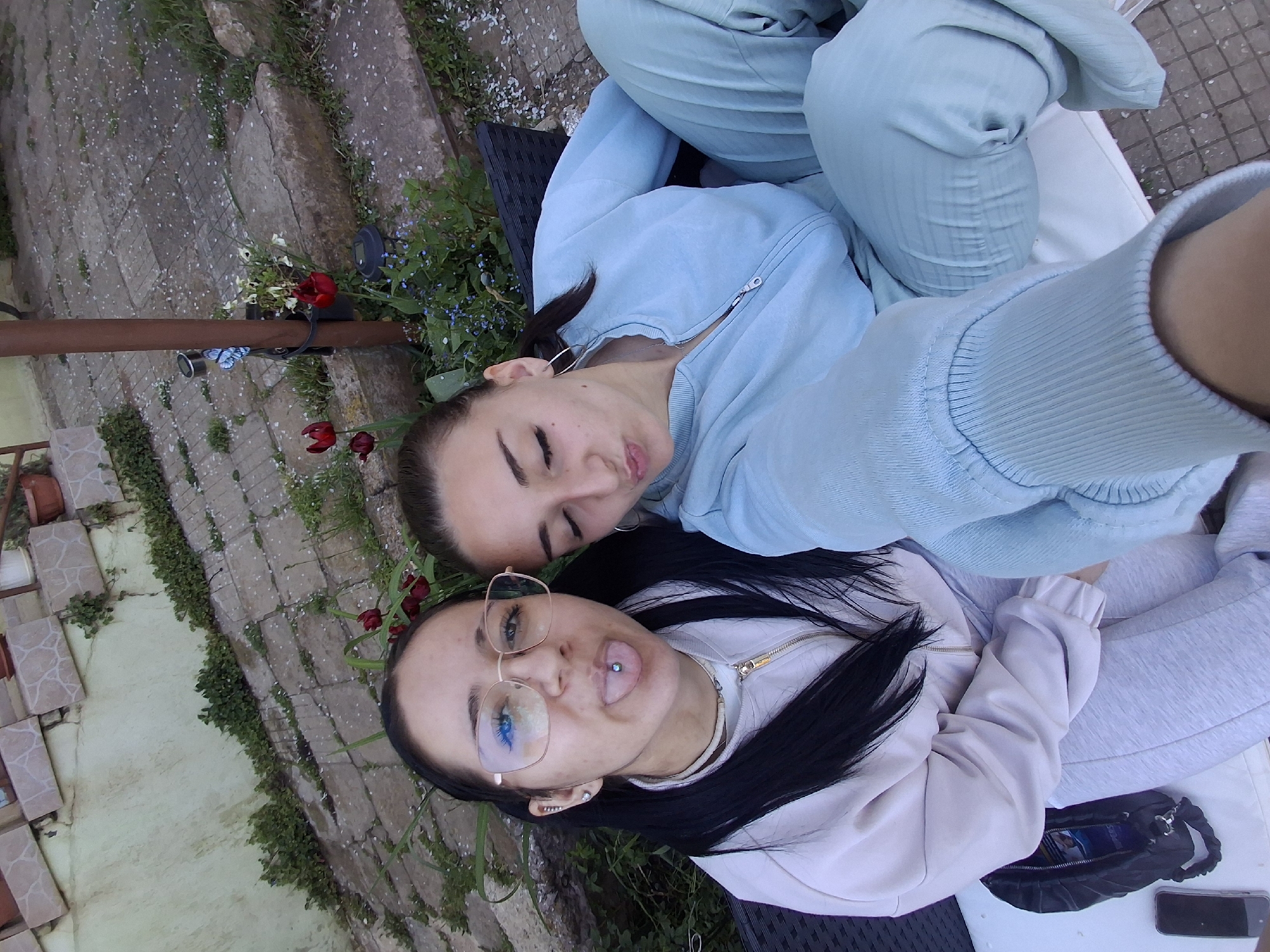 The image is a rotated overhead selfie featuring two young women relaxing outdoors in what appears to be a garden or patio setting in Buhovo, Bulgaria.
The woman positioned at the top of the frame has dark hair and is wearing a light blue or mint green hooded tracksuit. Her eyes are mostly closed, and she has a soft, relaxed smile.
The woman closer to the bottom of the frame also has dark hair and is wearing clear-framed glasses with blue-tinted lenses. She is dressed in a light-colored (possibly white or light pink) hoodie and grey sweatpants. She is playfully sticking out her tongue, which clearly displays a silver piercing. Next to her, a small black bag or pouch and a smartphone are visible on a white surface.
The background consists of a stone-paved path or patio, with areas of green grass and scattered small white petals, suggesting blossoms have fallen. Various plants and flowers, including red tulips, small blue flowers (possibly forget-me-nots), and white flowers, are visible in the surrounding greenery. A brown metal pole and a dark outdoor fixture (possibly a light or speaker) are also present. The scene suggests a casual and friendly outdoor gathering during daylight hours, with no specific time of day or weather conditions explicitly indicated beyond being suitable for outdoor leisure. No visible text is discernable in the image.