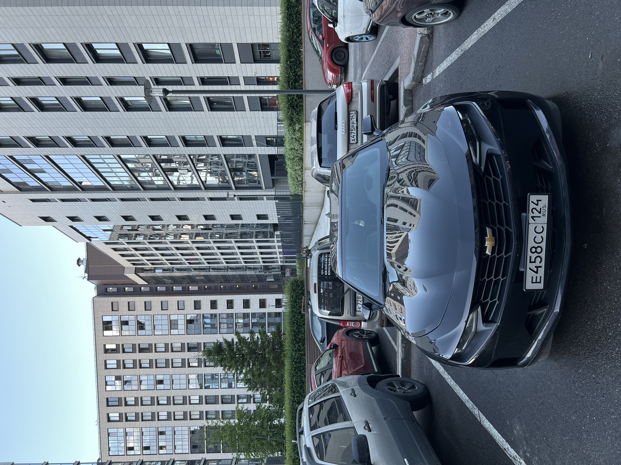 A black Chevrolet Camaro is parked in a parking lot in front of a modern apartment building in an unknown city in Russia. The car is the main subject, positioned centrally in the frame and facing the viewer. It is a two-door sports car with a sleek design. The license plate reads "E458CC 124 RUS".
Behind the Camaro, other vehicles are visible, including a silver Audi Q3, a red hatchback, a silver SUV, and a dark colored vehicle. The Audi Q3's license plate reads "A475PP 147". The parking lot surface is asphalt, marked with white lines delineating parking spaces.
The background features a large, contemporary apartment complex with many windows reflecting the sky and surrounding buildings. The architecture is characterized by a combination of light-colored facades and darker window frames, creating a geometric pattern. Green trees and hedges are planted in front of the buildings, adding a touch of nature to the urban landscape. The sky is partly cloudy. The overall scene suggests a residential area on a clear day.