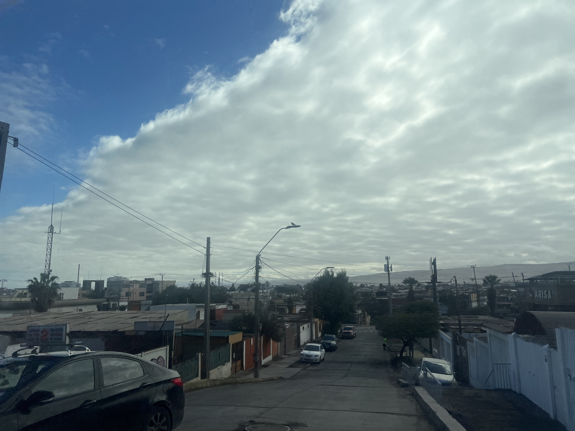 The image depicts a street scene in Arica, Chile, characterized by a mix of residential buildings and infrastructure under a partly cloudy sky. The perspective is from a vehicle, looking down a paved street that slopes slightly. On the left side of the street, a dark sedan is parked, partially visible. Further along, there are several houses with varying styles and colors, some featuring fences and gates. Utility poles with numerous wires crisscross the scene, indicating an urban or semi-urban environment.
Several cars are visible on the street: a white sedan, a dark SUV, and a red pickup truck further up the road. A person in a yellow vest is seen walking on the right side of the street near the pickup truck. In the background, more buildings, including some larger structures and what appears to be a commercial building with the name "ARISA" visible, are present. Palm trees are scattered throughout the landscape, and distant hills or mountains form the horizon. The overall atmosphere suggests a daytime scene in a moderately populated area of Arica.