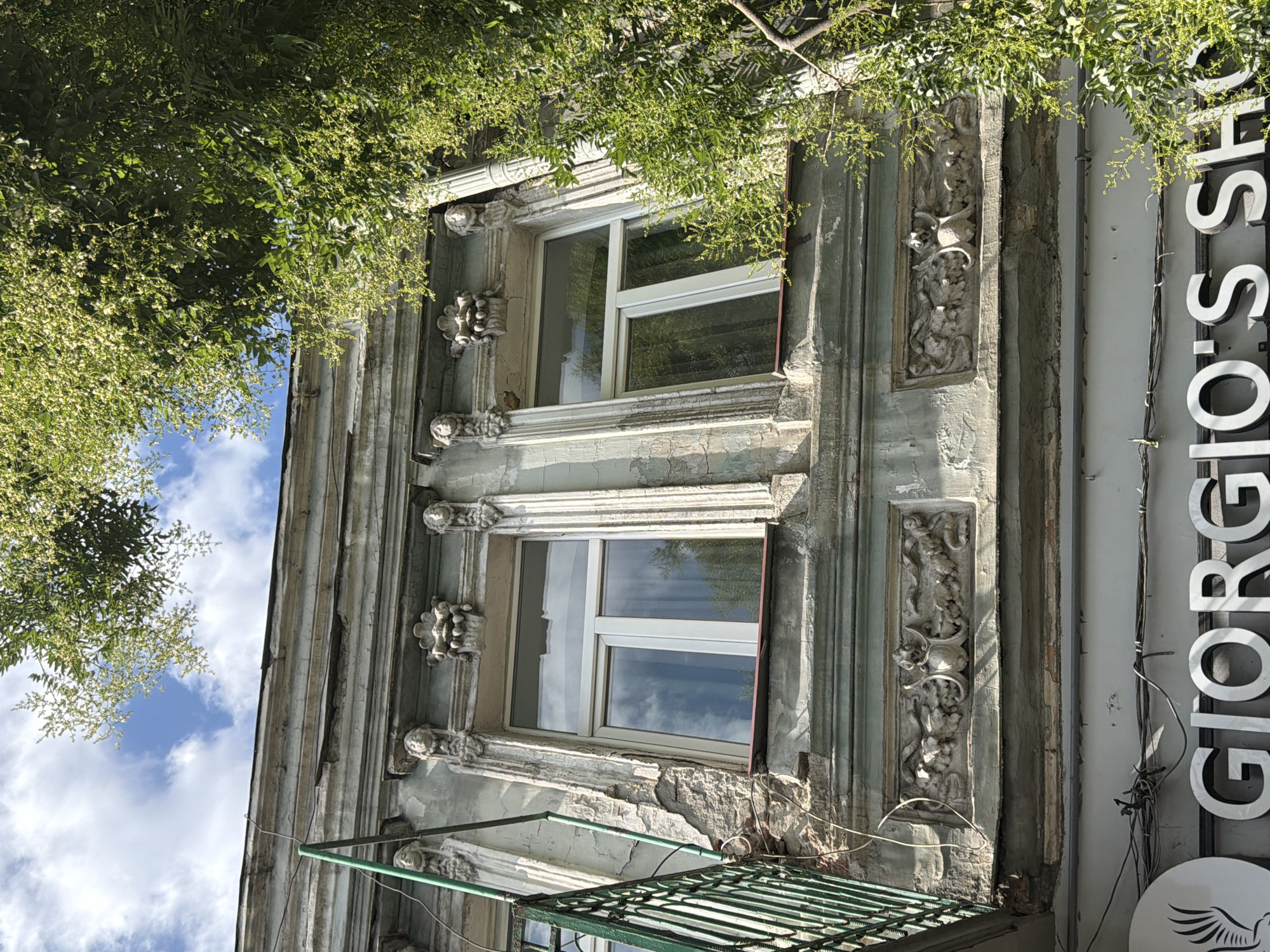 The image shows an upward view of the facade of an old building in Tbilisi, Georgia, with lush green trees partially obscuring the view. The building's architecture features ornate details, including carved faces above two white-framed windows, and a decorative frieze with mythological figures below. The facade shows signs of wear and tear, with peeling paint and crumbling plaster. In the lower part of the image, a sign for "GIORGIO'S SHOP" is visible, along with electrical wires. The sky is blue with scattered white clouds.
