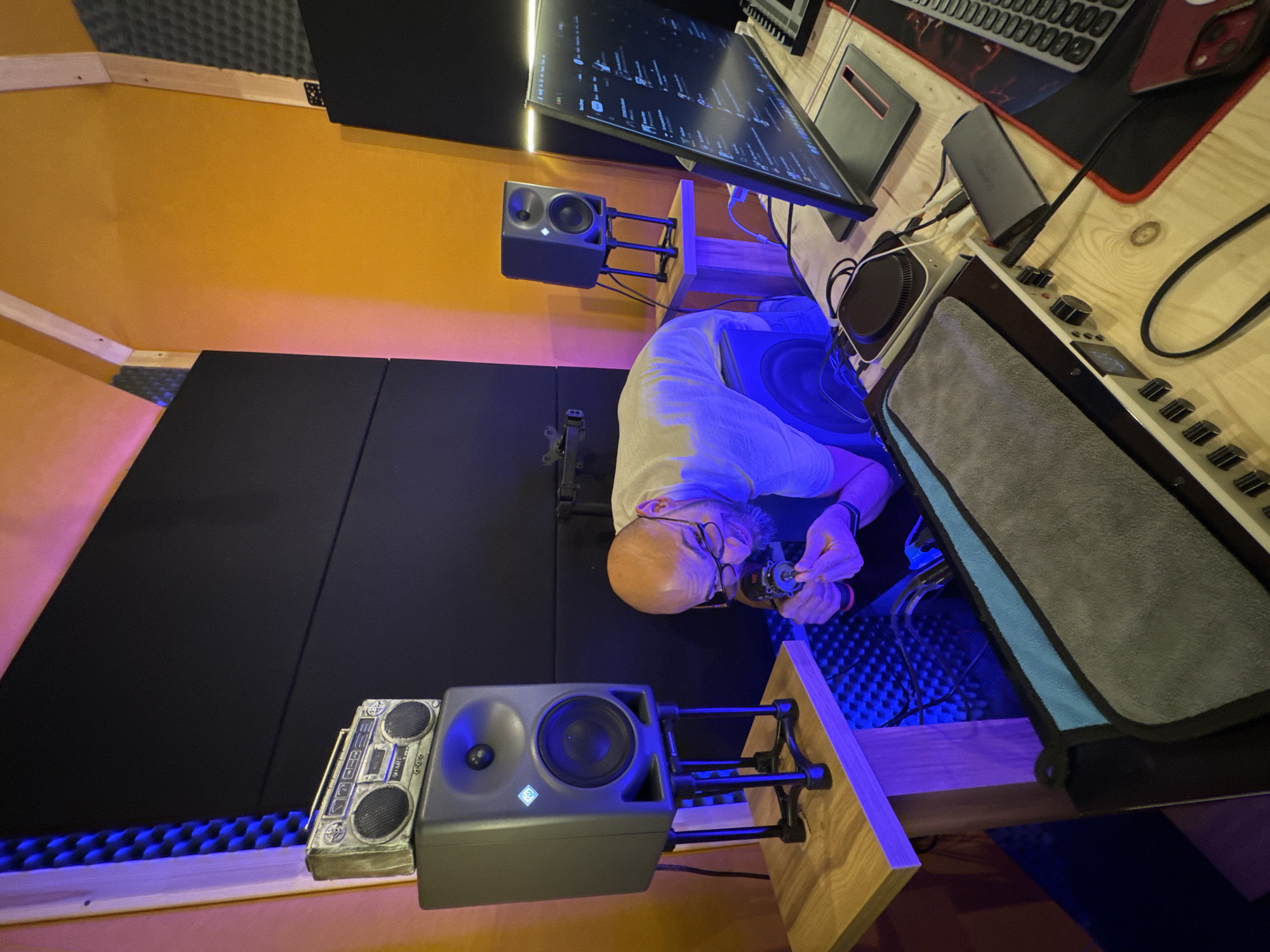 A man is shown working on a small object while in a recording studio. The man has short hair and is wearing glasses, a light-colored shirt, and a smartwatch on his left wrist. He appears to be holding the object in both hands and examining it closely. The recording studio features soundproofing panels on the walls, and several pieces of audio equipment, including studio monitors, a mixing board, a computer monitor, and a keyboard. A miniature boombox figurine sits near one of the speakers. A grey cloth with a blue lining is draped over a piece of equipment. The location is possibly a recording studio in Plovdiv, Bulgaria.