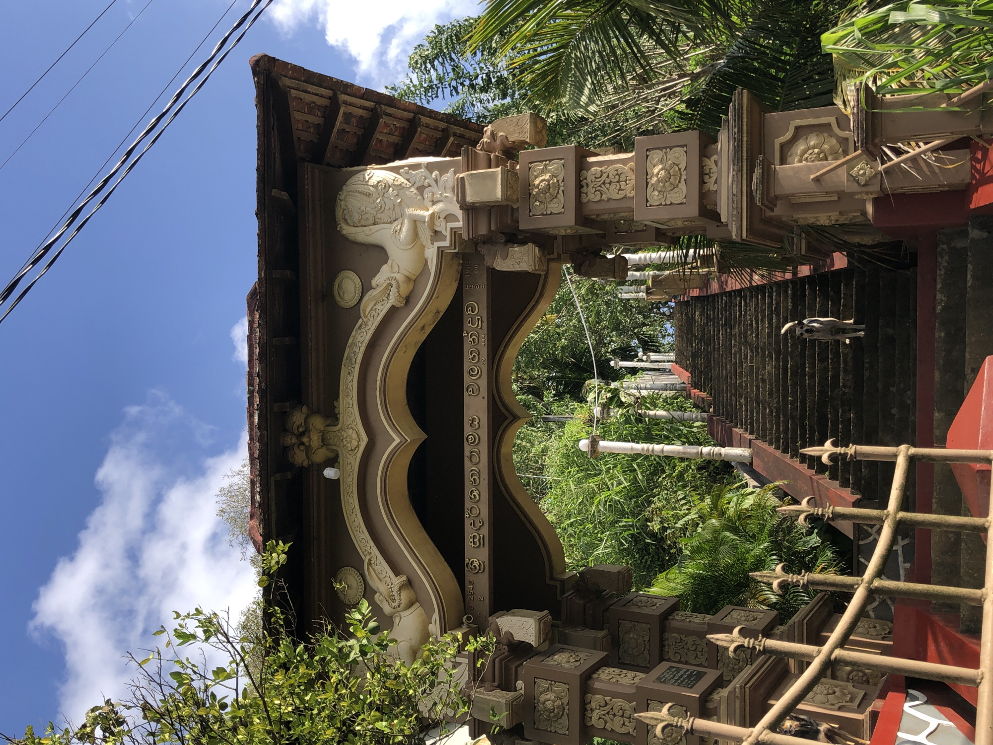 A medium shot captures a detailed view of an elaborately decorated archway leading to a long, stone staircase in Walahanduwa, Sri Lanka. The archway is adorned with intricate carvings, including what appear to be mythical creatures on either side of the central arch and decorative floral patterns on the supporting pillars. The roof of the archway is tiled with reddish-brown tiles, and in the background, lush green foliage and trees are visible. The sky above is a clear blue with a few wispy white clouds.
Dominating the foreground and midground of the image are the steps of a stone staircase, ascending towards an unseen destination. A small, light-colored dog is standing on the staircase, looking towards the camera. The staircase is flanked by a red-painted railing on the left and continues upwards into the dense greenery. The overall scene suggests a religious or cultural site, given the architectural style and the presence of the staircase leading upwards. The lighting indicates it is daytime, likely morning or afternoon, with sunlight casting shadows and highlighting the textures of the stone and carvings.