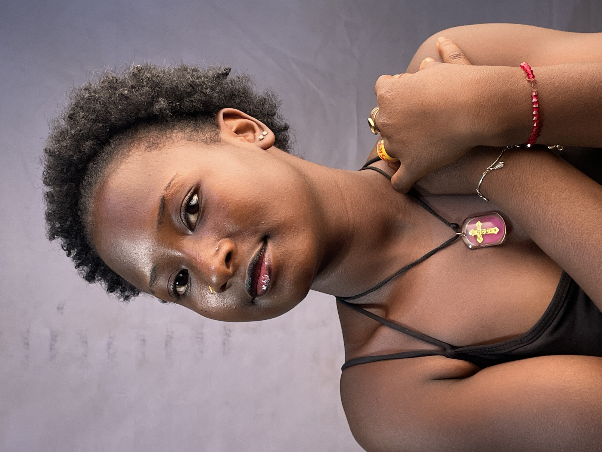 A medium shot features a dark-skinned woman with close-cropped curly black hair. She is gazing directly at the camera with a slight smile. She wears a black top with thin straps, a nose ring, and a pendant necklace with a pink and gold cross. On her right hand, a red beaded bracelet and multiple rings adorn her fingers. Her hands are clasped together near her neck. The background is a smooth, gray gradient. There is no indication of a location in the city of Jos, Nigeria, or any external location, or context. The image appears to be a studio portrait.