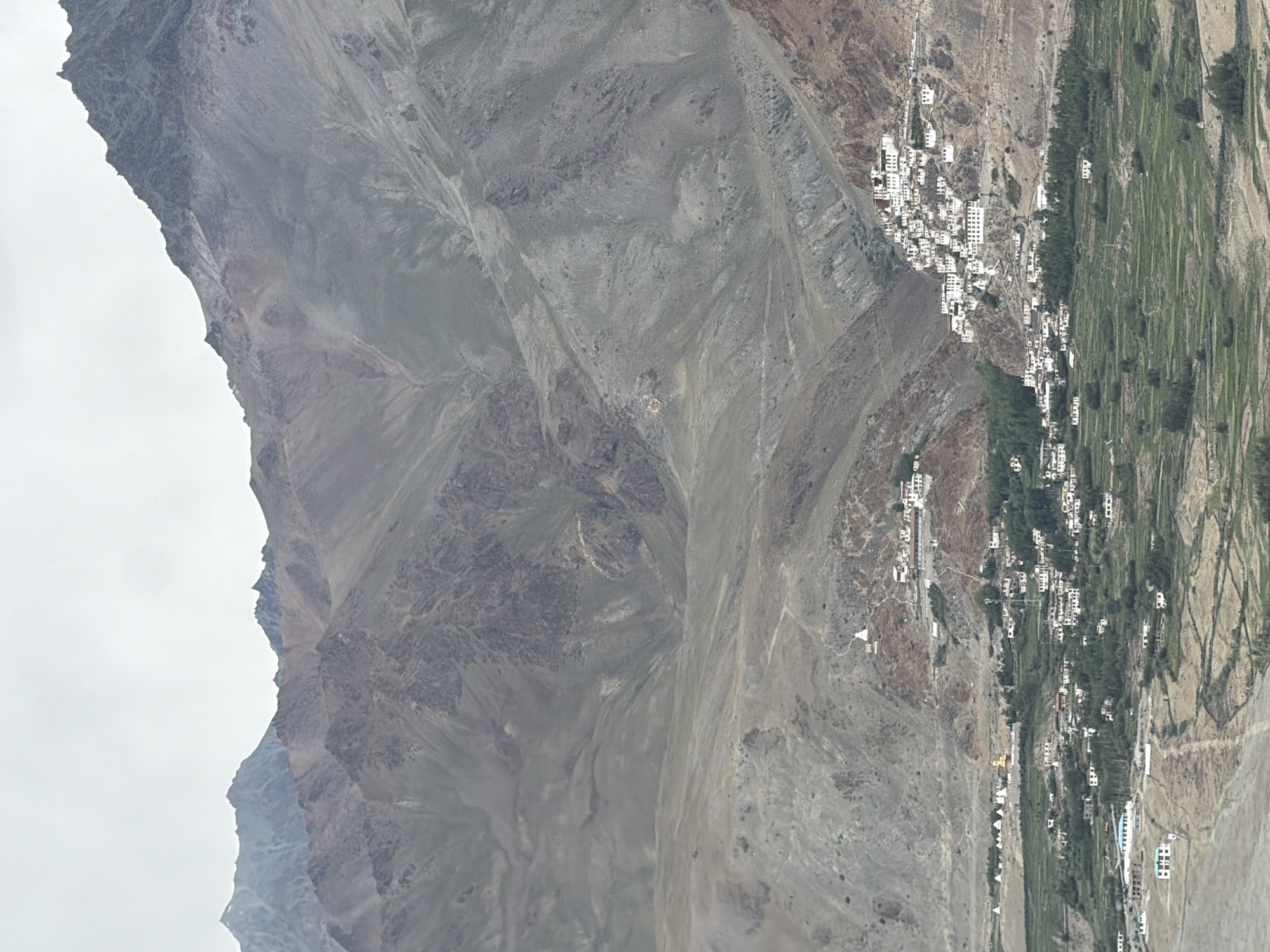 The image depicts a mountainous landscape in Pibiting, India. The scene features a cluster of white buildings with dark roofs cascading down the side of a steep, rocky mountain. These structures appear to be part of a settlement or monastery. Below the mountain, a verdant valley is visible, with terraced fields indicating agricultural activity. A road winds through the valley, connecting various scattered buildings. The sky is overcast, casting a diffused light on the scene. The overall impression is one of a remote, high-altitude location with a distinctive architectural style integrated into the natural environment.
