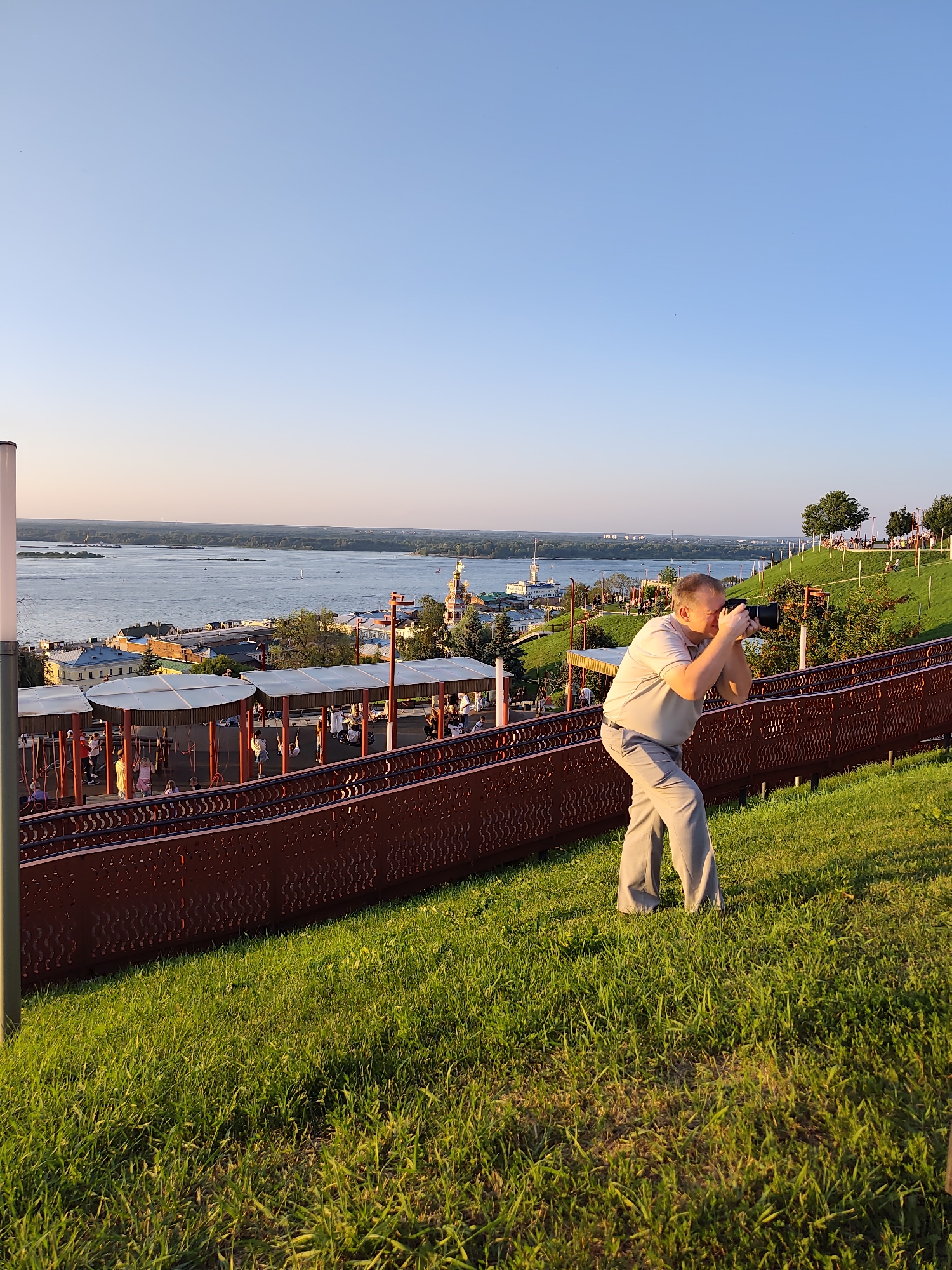 The image captures a photographer, a middle-aged man wearing a light-colored polo shirt and grey trousers, actively taking a picture with a professional camera. He is positioned on a grassy slope in a park overlooking a wide river. The landscape suggests the city of Nizhny Novgorod, Russia, with its distinctive architecture and the confluence of rivers visible in the background. Below the photographer, a red metal railing separates the grassy area from a covered walkway or public space where several people, including children, can be seen. The sky is clear and blue, indicating it is likely late afternoon or early evening due to the warm, golden light illuminating the scene. The overall impression is one of leisure and sightseeing in a scenic urban environment.