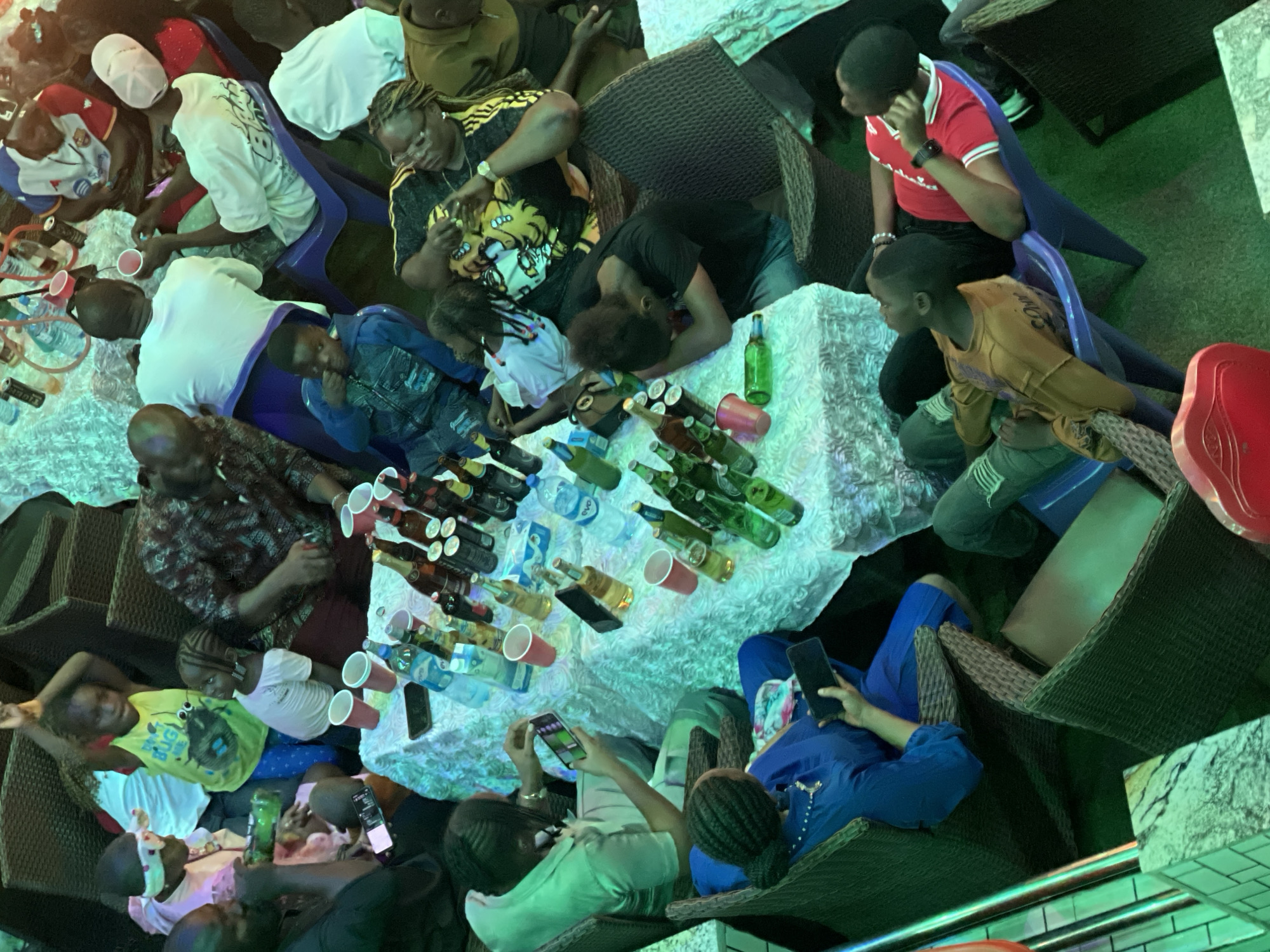 The image depicts an overhead view of a gathering of people around a table heavily laden with bottles, cans, and red plastic cups, potentially at a bar or recreational establishment. A large group of people are interacting around a white textured table. Some are seated in plastic chairs while others are in rattan-style armchairs. Some individuals are using mobile phones. The location is presumed to be Obosi, Nigeria.