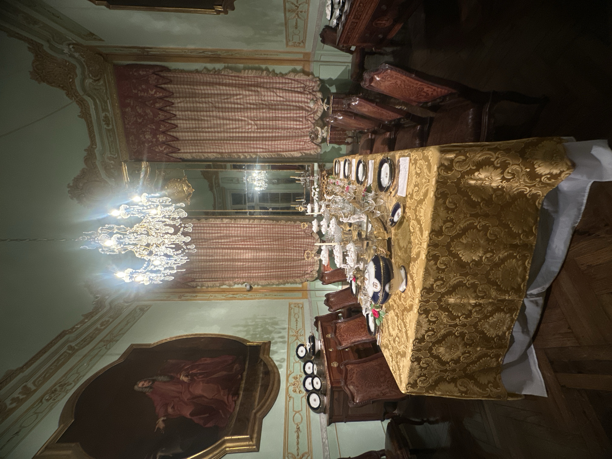 The image depicts a dining room in a historical building, likely a museum or palace in Genoa, Italy, given the ornate decor. The room features a large chandelier, a long table set with fine china and glassware, and a painting in an elaborate frame. The walls are decorated with intricate moldings and designs, and there are heavy drapes at the windows. Several chairs are arranged around the table, suggesting a formal dining setting. The scene is dimly lit.