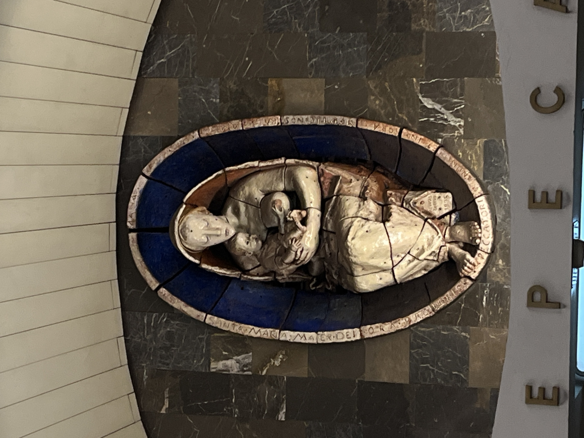 The image shows a relief sculpture of a woman holding a baby, mounted on a dark, marbled wall. The sculpture is framed by a blue and brown oval border. The figures are rendered in a stylized manner with cracked, textured surfaces. Inscriptions are visible around the border of the sculpture. Below the sculpture, a white band with large, gold-colored letters spelling "EPECA" is visible. The overall setting appears to be an interior, possibly a public space given the architectural elements and lettering. The style of the sculpture and the lettering suggests it could be located in Moscow, Russia, possibly in a metro station or a similar public building.