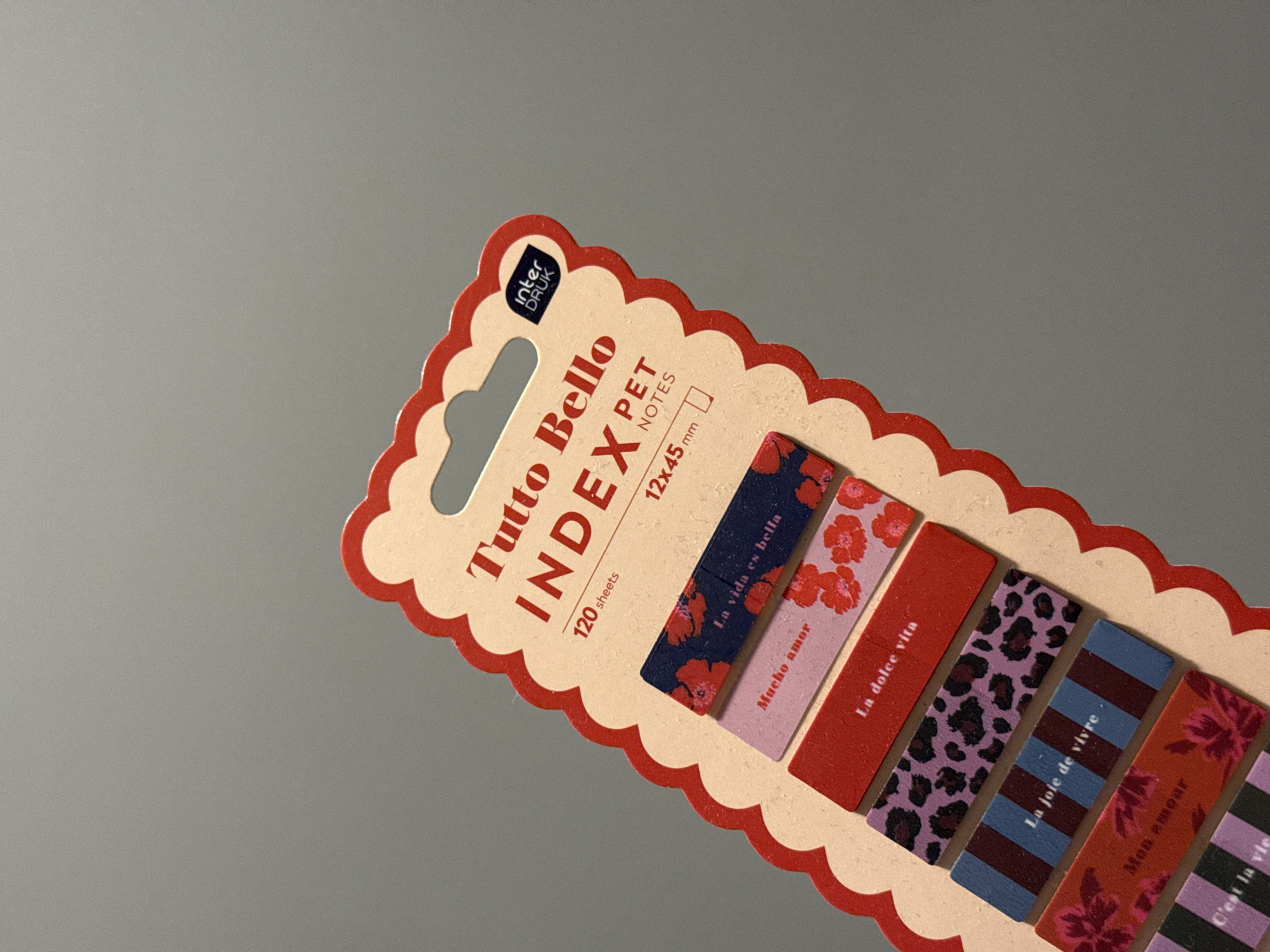 The image shows a product display card for a set of index tabs. The card is red and cream colored with a scalloped edge. The product name, "TUTTO BELLO INDEX PET NOTES," is printed in large red letters. Below this, it states "120 sheets" and "12x45 mm."
Attached to the card are seven individual index tabs, each with a different pattern and text in a foreign language. The tabs are arranged vertically and partially overlap each other. The patterns include floral designs in red and pink, a navy blue floral pattern, a leopard print in purple and black, a striped pattern in blue and burgundy, and a striped pattern in pink and purple. The text on the tabs appears to be in Italian and French: "La vita es bella," "Mucho amor," "La dolce vita," "La joie de vivre," "Mon amour," and "C'est la vie." The tabs are made of a matte material. A small circular logo with "inter DRUK" is visible at the top right of the card.
The background of the image is a plain, neutral gray surface, suggesting the product is placed on a table or shelf. There are no people or other objects visible in the scene. The lighting is even, with no strong shadows. The image is a product shot, likely for e-commerce or promotional purposes.
This image does not contain any information that would allow for the determination of its location in the city of Łódź, Poland.