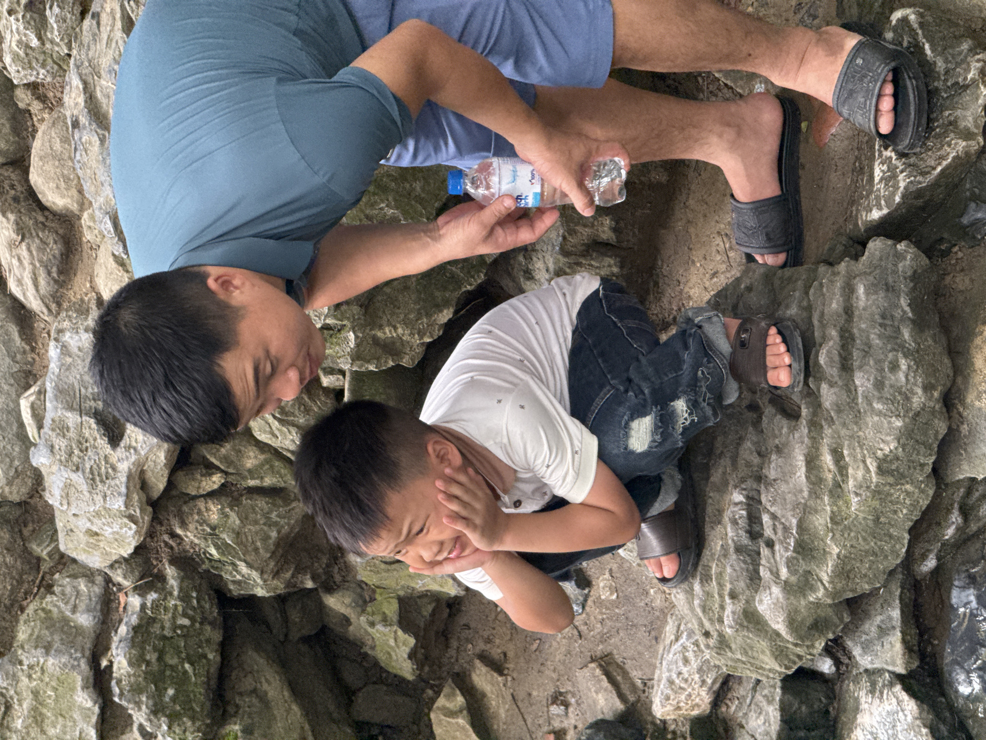 In the city of Cẩm Tú Commune, Vietnam, a boy in a white t-shirt and ripped jeans is squatting on a rock, his hands on his cheeks as he looks to the side. He is wearing brown sandals. An adult male, wearing a teal polo shirt and blue shorts, is leaning over and holding a bottle of water. He is wearing black slide sandals. The two individuals are positioned near a rocky embankment with sparse vegetation.