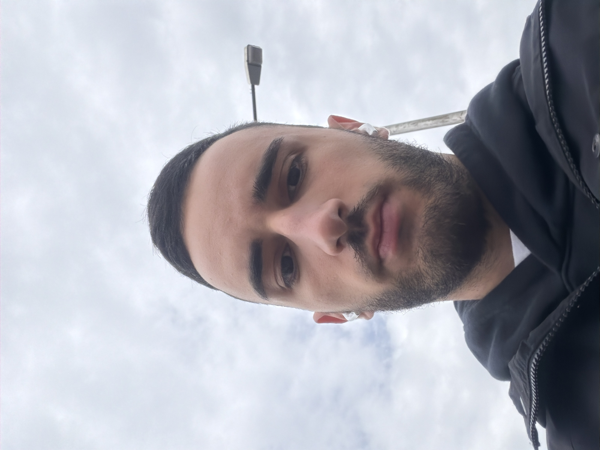 A man with short dark hair, a beard, and brown eyes is wearing a black hooded jacket and white wireless earbuds. He is looking directly at the camera. Behind him is a cloudy sky and a street lamp. The location is unknown and cannot be determined from the image.