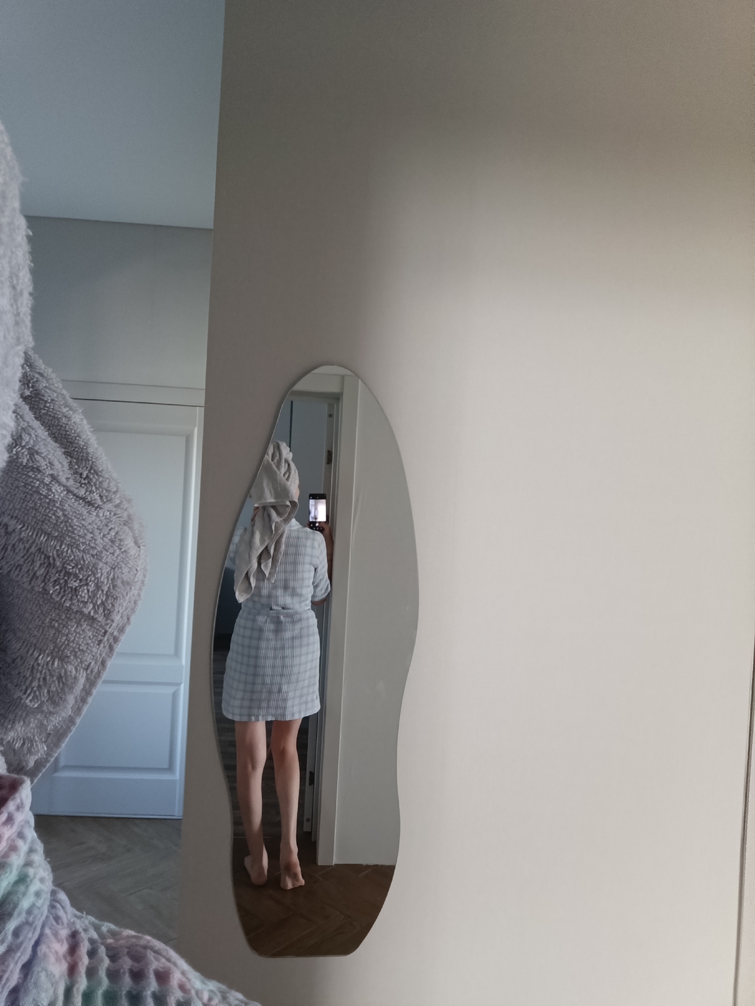 This image depicts an indoor scene in Chaikovsky, Russia. The primary subject is a person, seen from behind, reflected in an irregularly shaped wall mirror. The individual is wearing a light-colored, possibly blue and white plaid, short-sleeved garment extending to mid-thigh, and a light-colored towel is wrapped around their head. They are barefoot, standing on a wooden floor with a herringbone or parquet pattern. The person is holding a black smartphone towards the mirror, with the screen or flash illuminated, indicating the act of taking a photograph of their reflection. The background in the mirror shows a light-colored door or wall. The foreground, outside the mirror's reflection, includes a fluffy, gray towel or similar textile object partially visible on the left, beneath which a multicolored knitted item in pastel shades of blue, pink, and green is visible. The wall on which the mirror is mounted is a plain, light-colored surface.