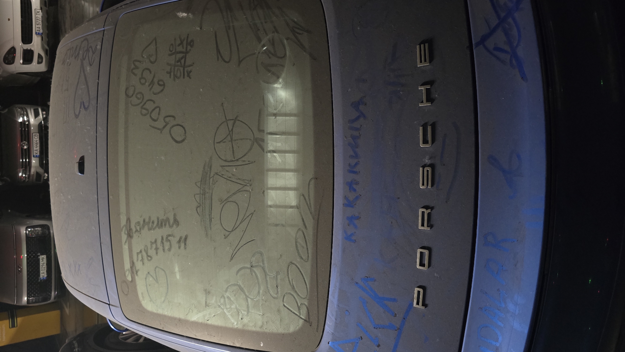 An overhead-angle image depicts the rear of a dark blue/purple Porsche vehicle, heavily covered in dust and marked with various writings and drawings, positioned within what appears to be an underground parking garage in Kyiv, Ukraine.
The primary subject is the dust-covered Porsche. The vehicle's rear window is significantly obscured by a thick layer of dust, into which multiple patterns, numbers, and words have been etched. Visible dust markings include:
* On the left side, "arina" (likely Cyrillic "Арина") is written vertically, followed by the numbers "787511".
* In the center-bottom, "BoooM" is written in large letters.
* Above "BoooM", a stylized word resembling "NeMo" is present, along with other overlapping scribbles.
* To the left of "NeMo", "2020" is faintly visible.
* On the right side, the numbers "050960" are written vertically, with "6493" below them.
* Below "6493", a tic-tac-toe grid with 'X' and 'O' marks is drawn.
* A heart symbol and other indistinct scribbles are also visible on the window.
* On the roof, just above the rear window, the letters "XXX" are faintly visible in the dust.
Below the rear window, on the vehicle's trunk lid, the embossed "PORSCHE" emblem is centered. This area of the vehicle, also dusty, features additional graffiti applied with blue spray paint. A prominent Cyrillic word, "КАКАКИЩА" (Kakakisha), is written above the Porsche emblem. Further blue spray paint scribbles are present on both sides of the trunk lid and extending onto the rear bumper, with one discernible partial word appearing to be "DALAR".
In the background, other parked vehicles are visible, confirming the location as a parking facility. From left to right:
* A silver Hyundai Staria with Ukrainian license plate "KA 0296 MK".
* A silver Volkswagen (likely a Tiguan) with Ukrainian license plate "KA 4444 CB".
* A white Porsche Cayenne with Ukrainian license plate "AA 5777 TK".
All license plates indicate registration in Kyiv (KA) or Kyiv Oblast (AA), affirming the location as Kyiv, Ukraine.
The environment is an indoor, artificially lit parking garage, characterized by concrete walls and pillars, and yellow parking space delineations. The lighting consists of overhead fluorescent fixtures, casting reflections on the rear window of the Porsche. The overall condition of the Porsche suggests it has been stationary for an extended period, accumulating significant dust.